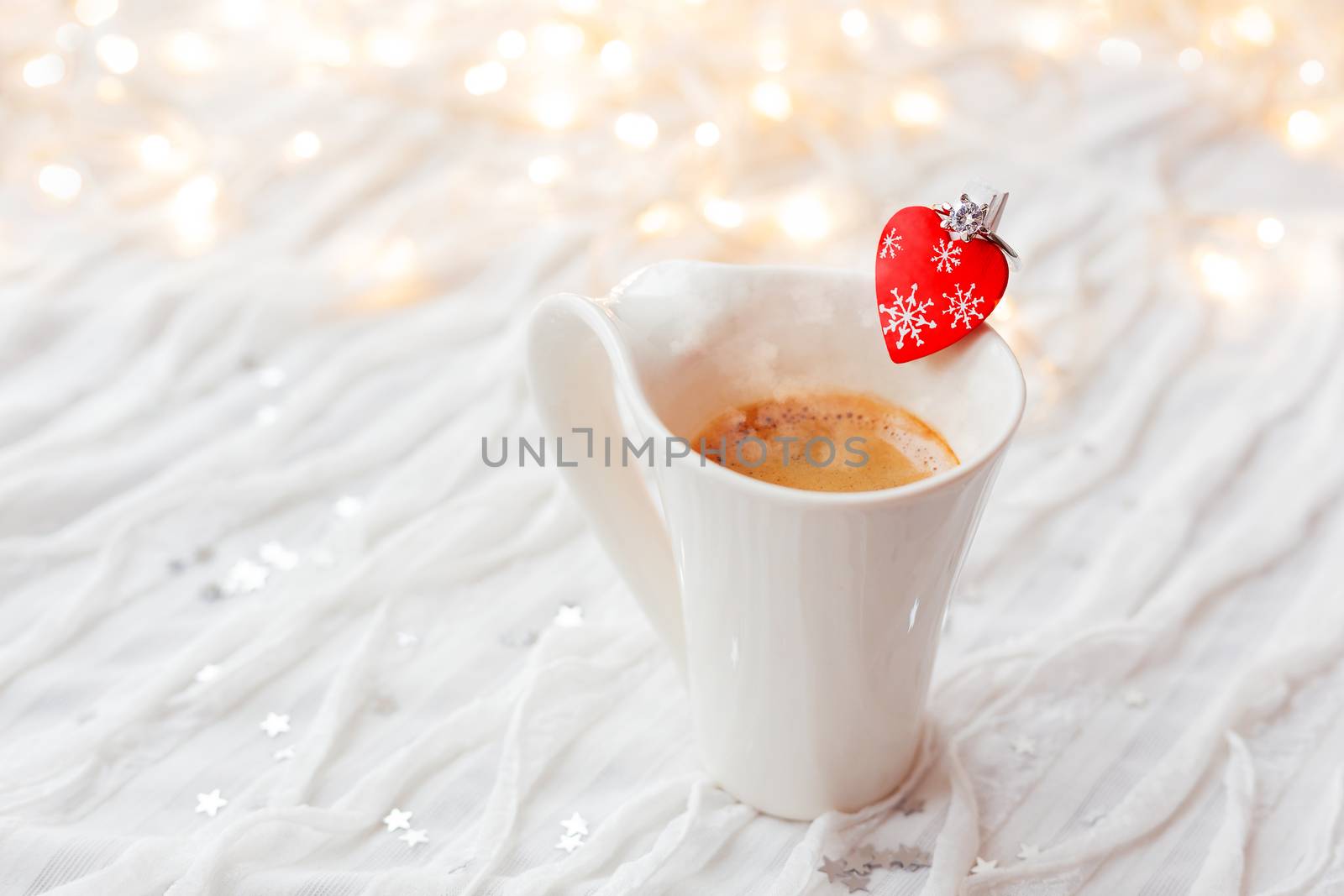 White cup of hot coffee with decorative heart and engagement diamond ring, symbol of love and marriage. Valentine's day background with lights and decorations.