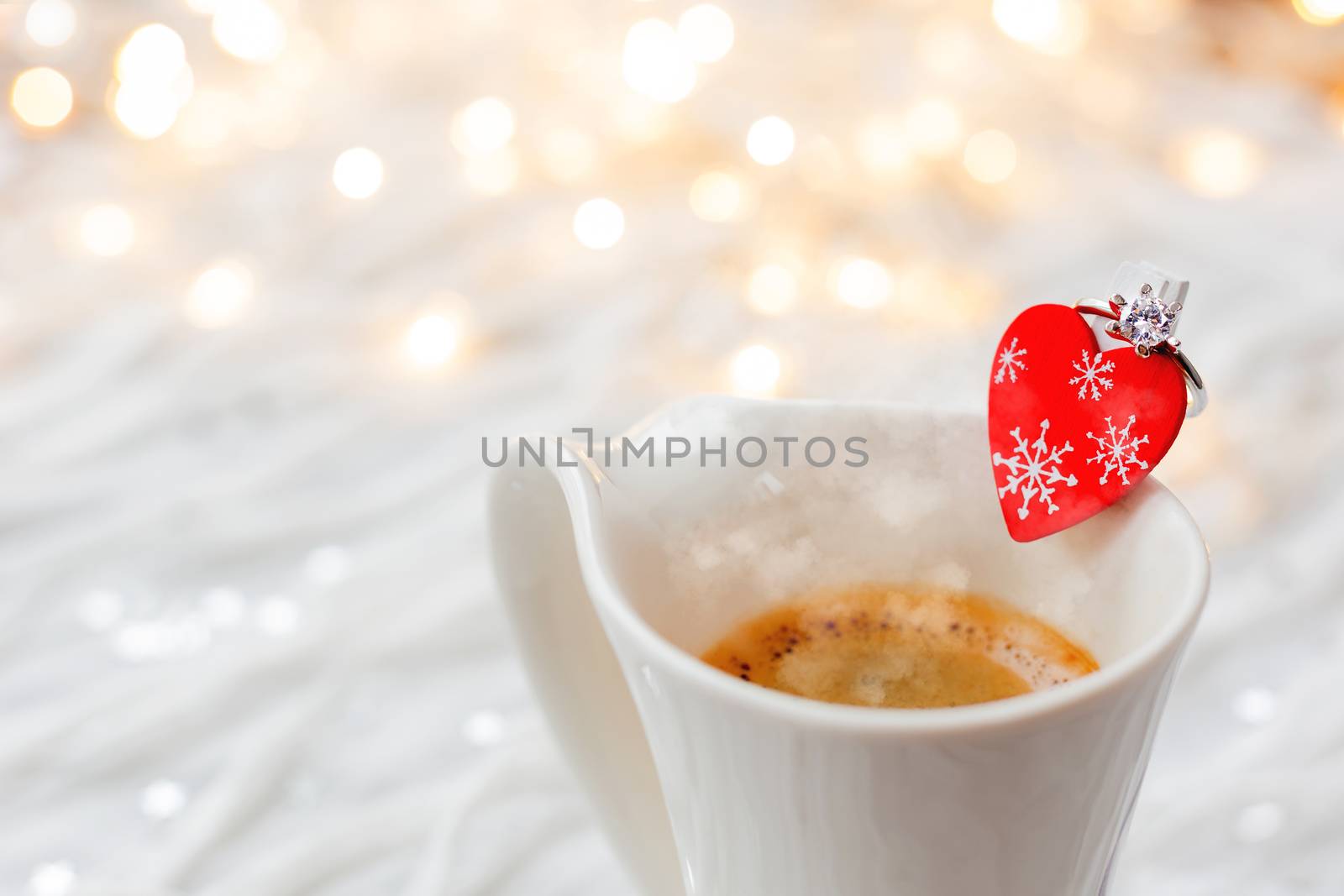 White cup of hot coffee with decorative heart and engagement diamond ring, symbol of love and marriage. Valentine's day background with lights and decorations.