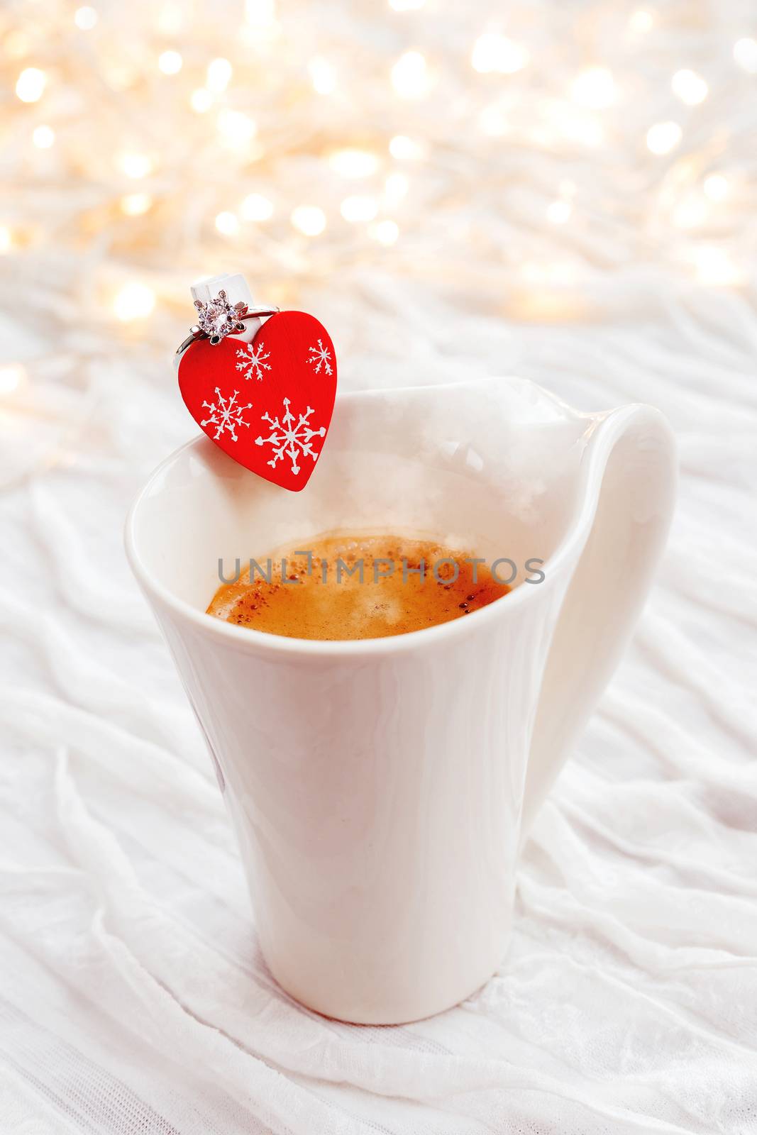 White cup of hot coffee with decorative heart and engagement diamond ring, symbol of love and marriage. Valentine's day background with lights and decorations.