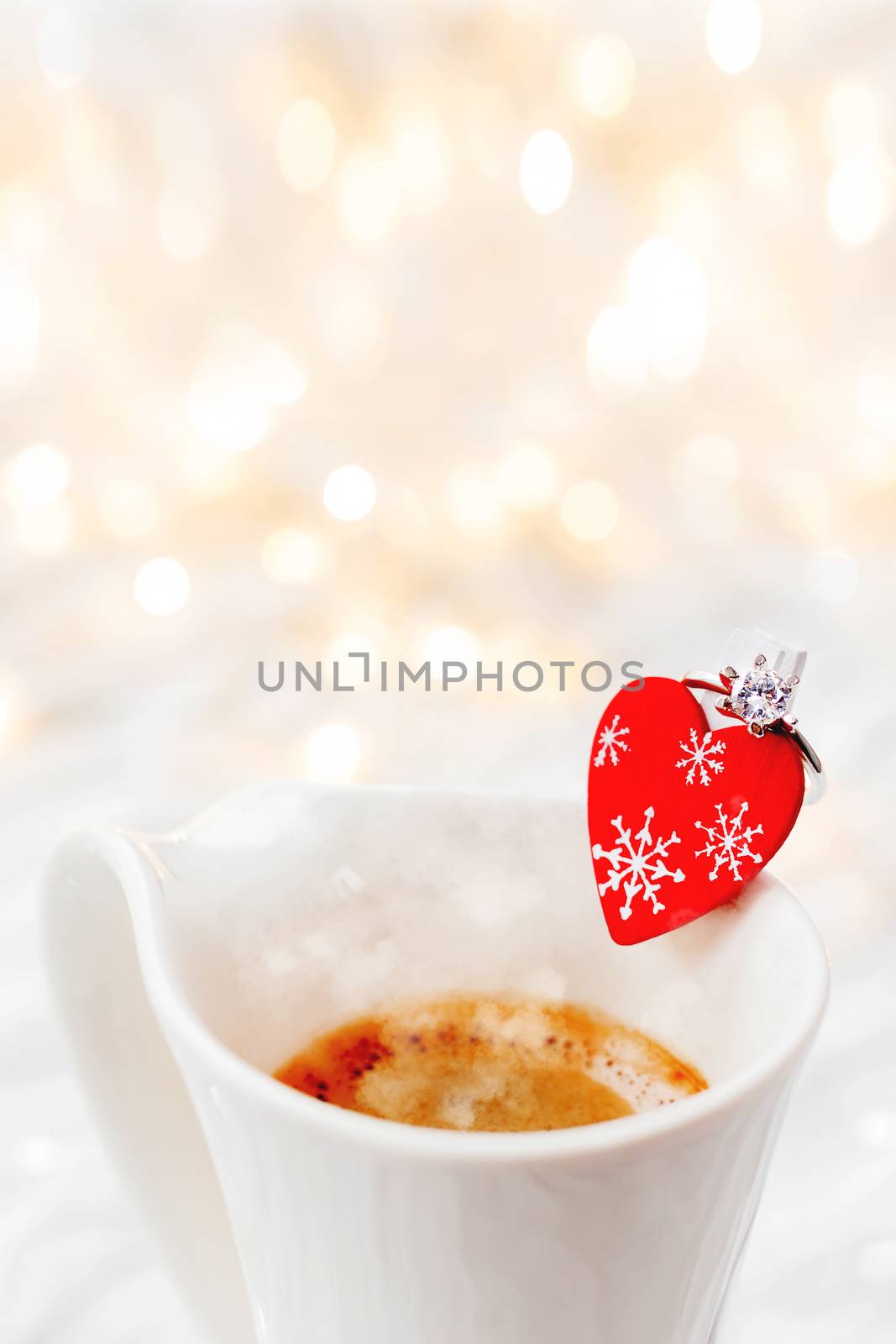 White cup of hot coffee with decorative heart and engagement diamond ring, symbol of love and marriage. Valentine's day background with lights and decorations.