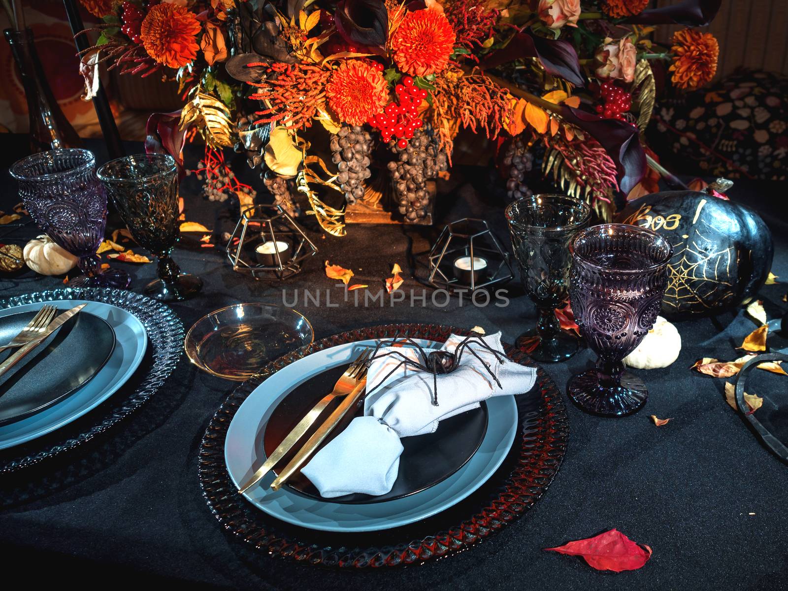 Table served in Halloween style. Bouquet with red flowers, decorative spiders, pumpkins with lettering and gothic vine glasses on black tablecloth. Holiday decorations.