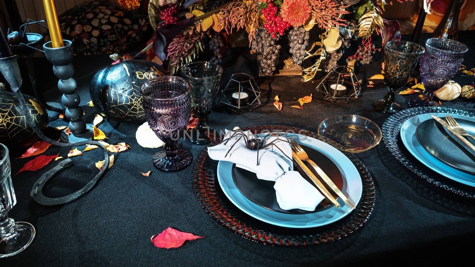 Table served in Halloween style. Bouquet with red flowers, decorative spiders, pumpkins with lettering and gothic vine glasses on black tablecloth. Holiday decorations. Place for text.