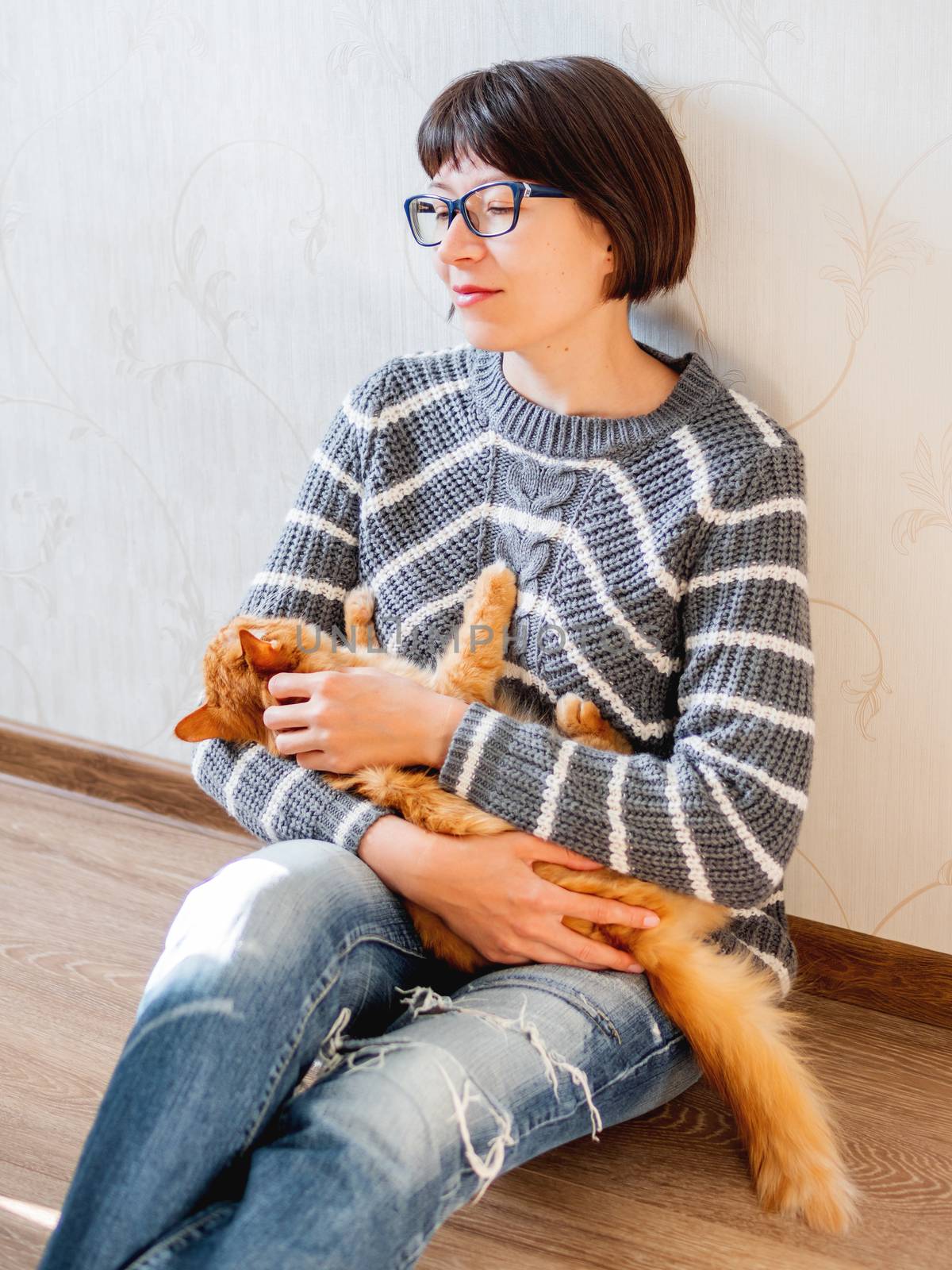 Cute ginger cat dozing on woman knees. Smiling woman in torn jeans stroking her fluffy pet. Cozy home.