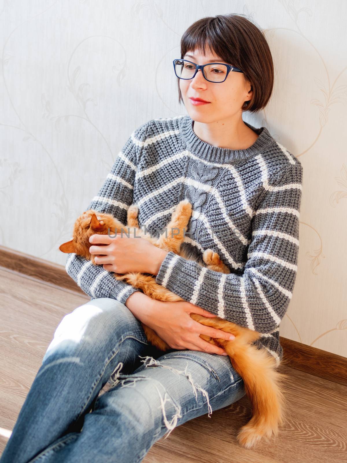 Cute ginger cat dozing on woman knees. Smiling woman in torn jeans stroking her fluffy pet. Cozy home.