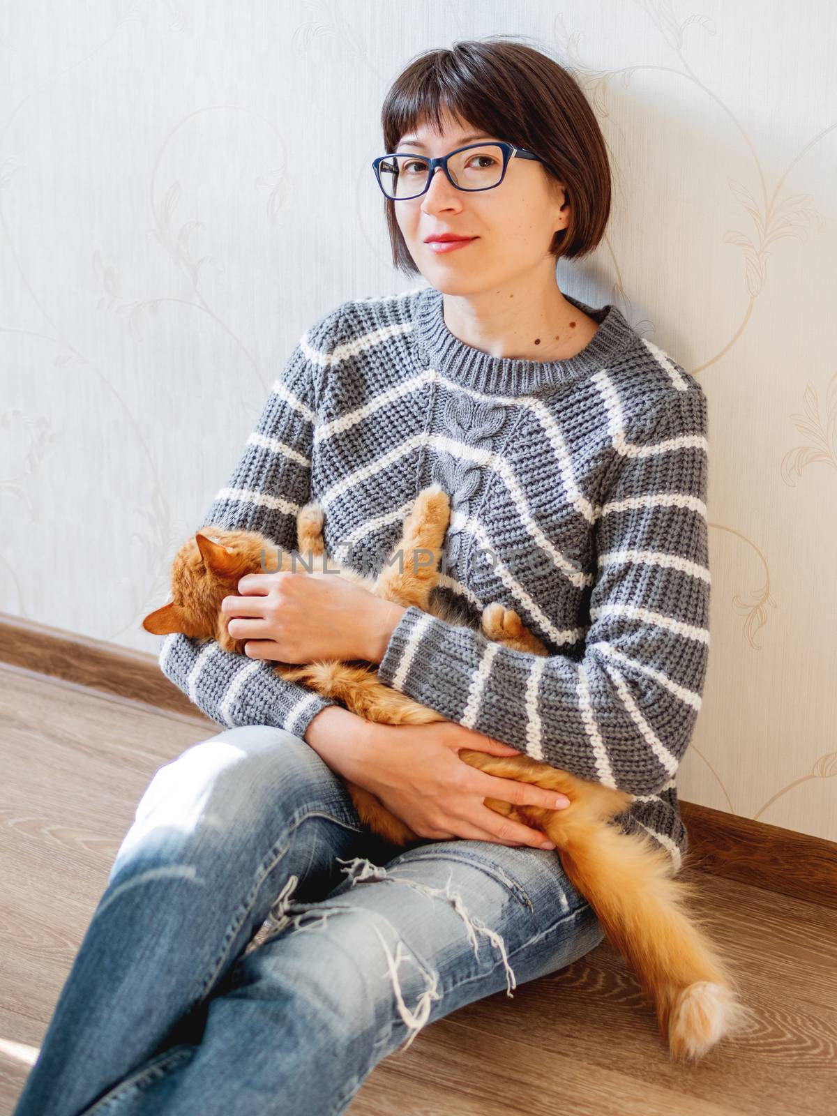 Cute ginger cat dozing on woman knees. Smiling woman in torn jeans stroking her fluffy pet. Cozy home.