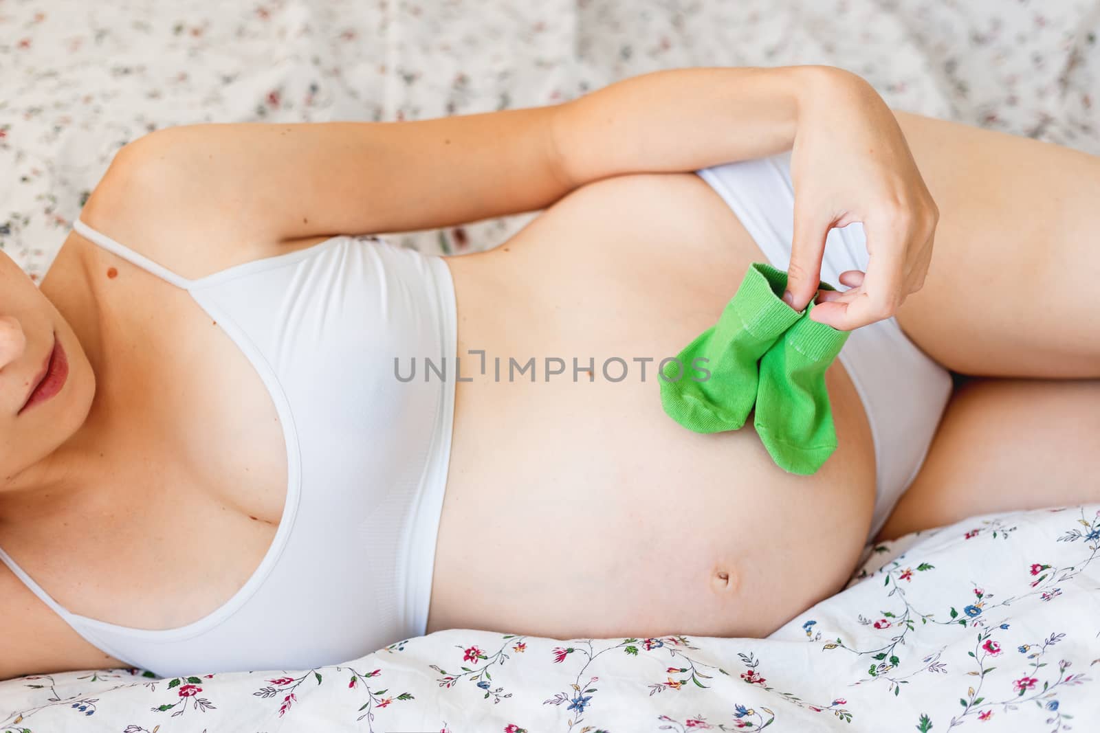 Pregnant woman in white underwear lying in bed with small baby's socks. Young woman expecting a baby.