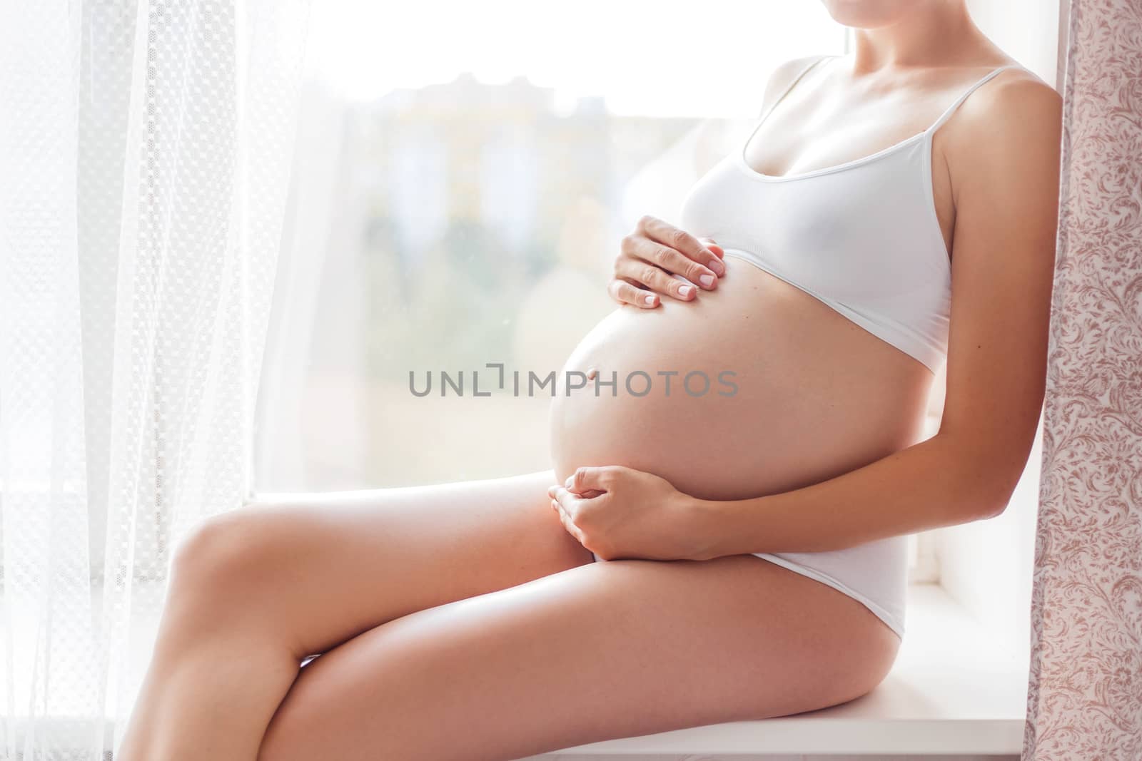 Pregnant woman in white underwear sitting on window sill. Young woman expecting a baby. Cozy happy background in sunny morning.