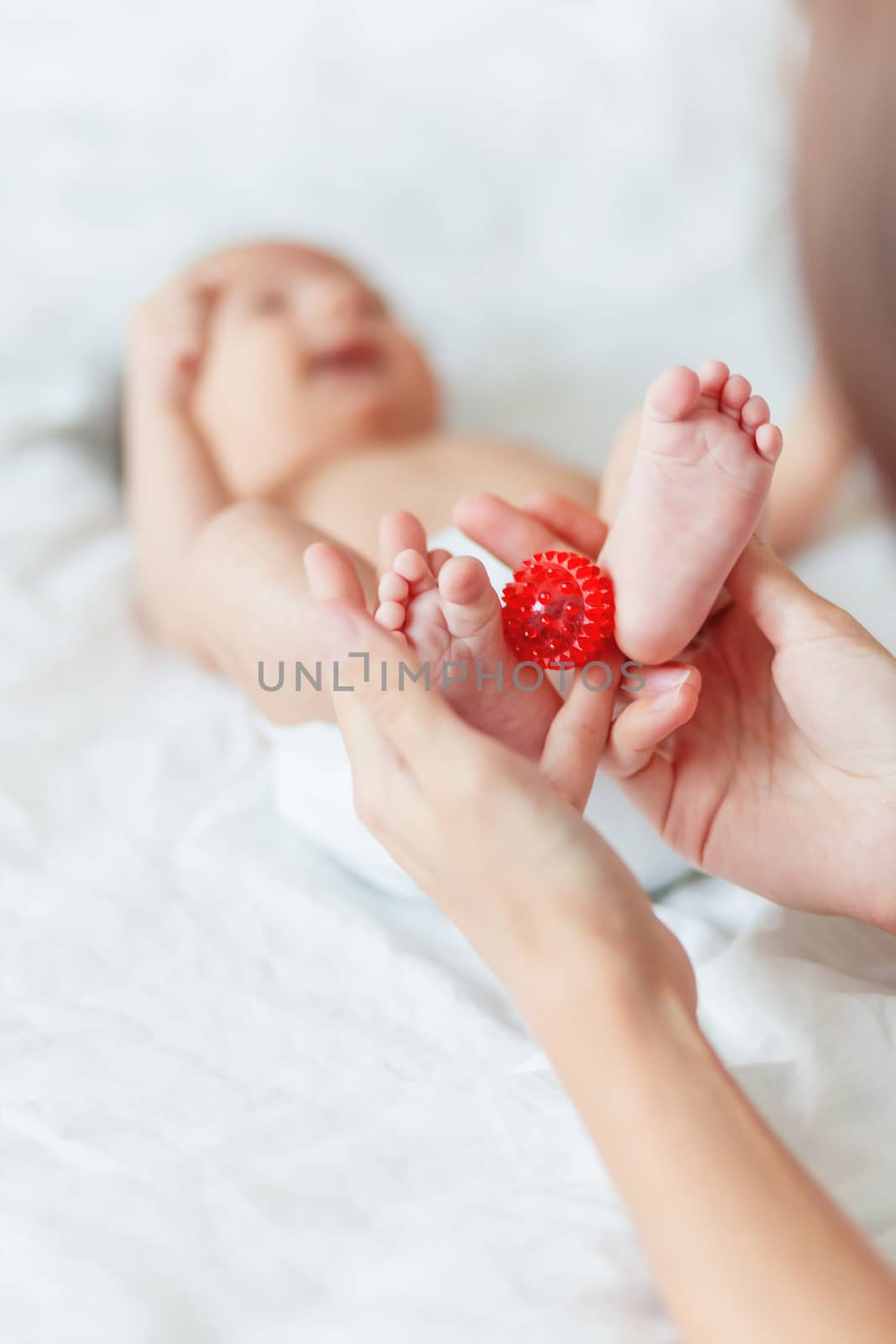 Mother holds newborn baby's feet. Tiny fingers and red massage ball in woman's hand. Cozy morning at home.