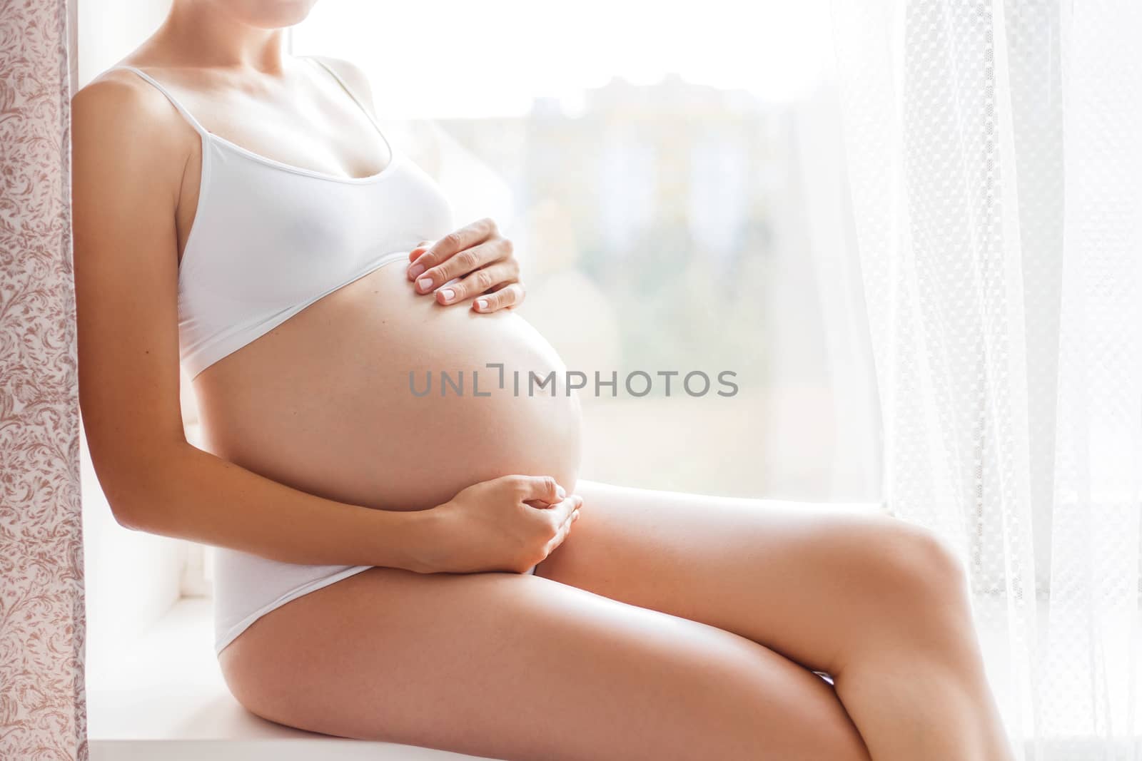 Pregnant woman in white underwear sitting on window sill. Young woman expecting a baby. Cozy happy background in sunny morning.