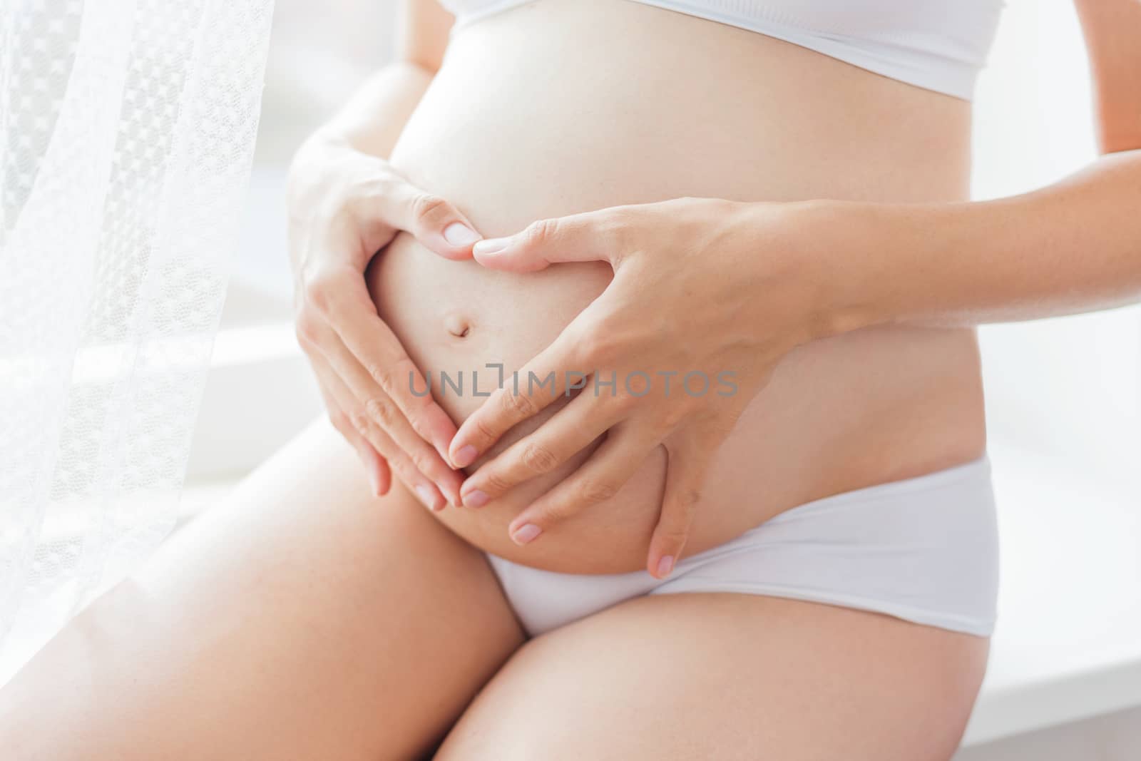 Pregnant woman in white underwear. Young woman expecting a baby. Future mother makes heart gesture - symbol of love.