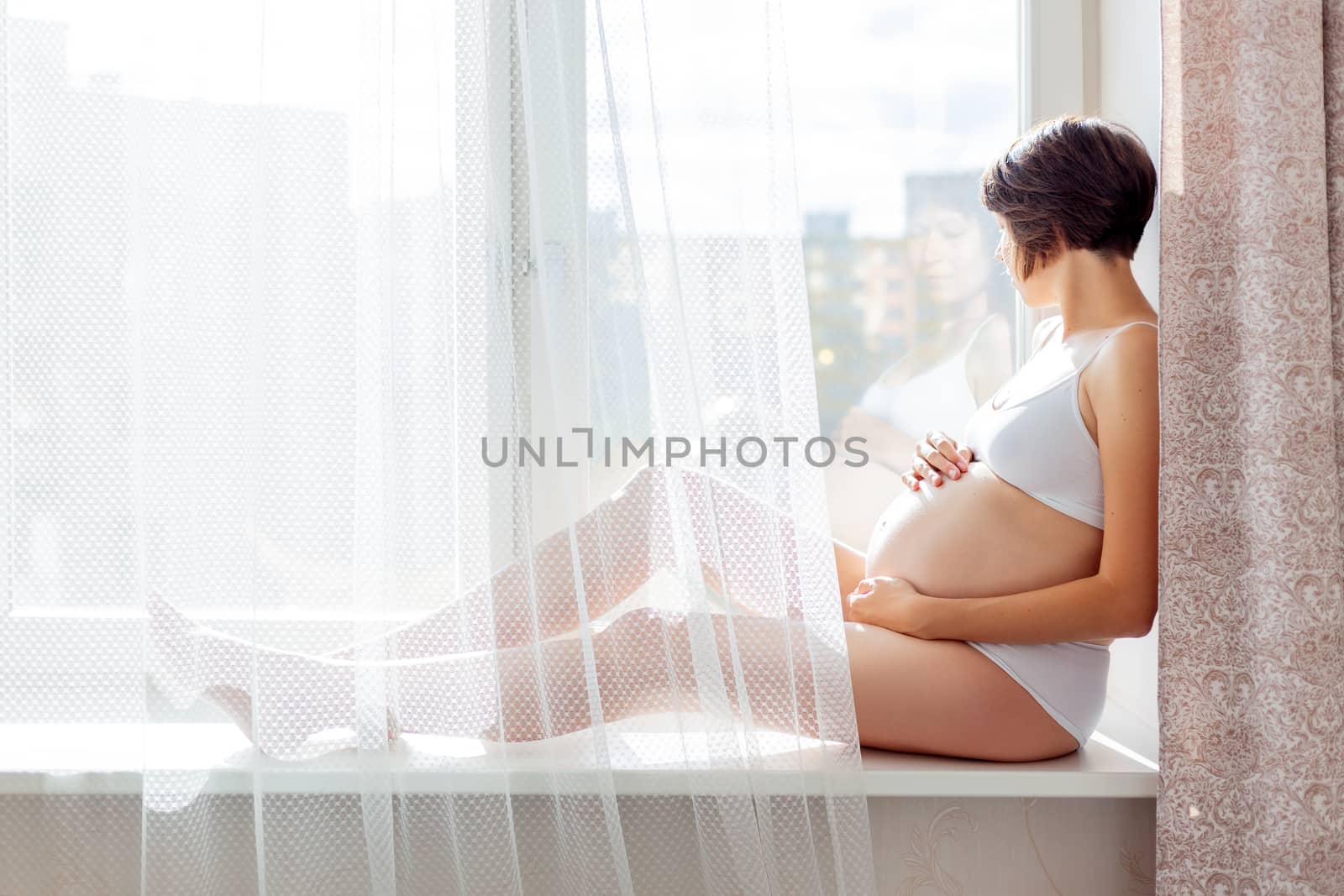 Pregnant woman in white underwear sitting on window sill. Young woman expecting a baby. Cozy happy background in sunny morning.