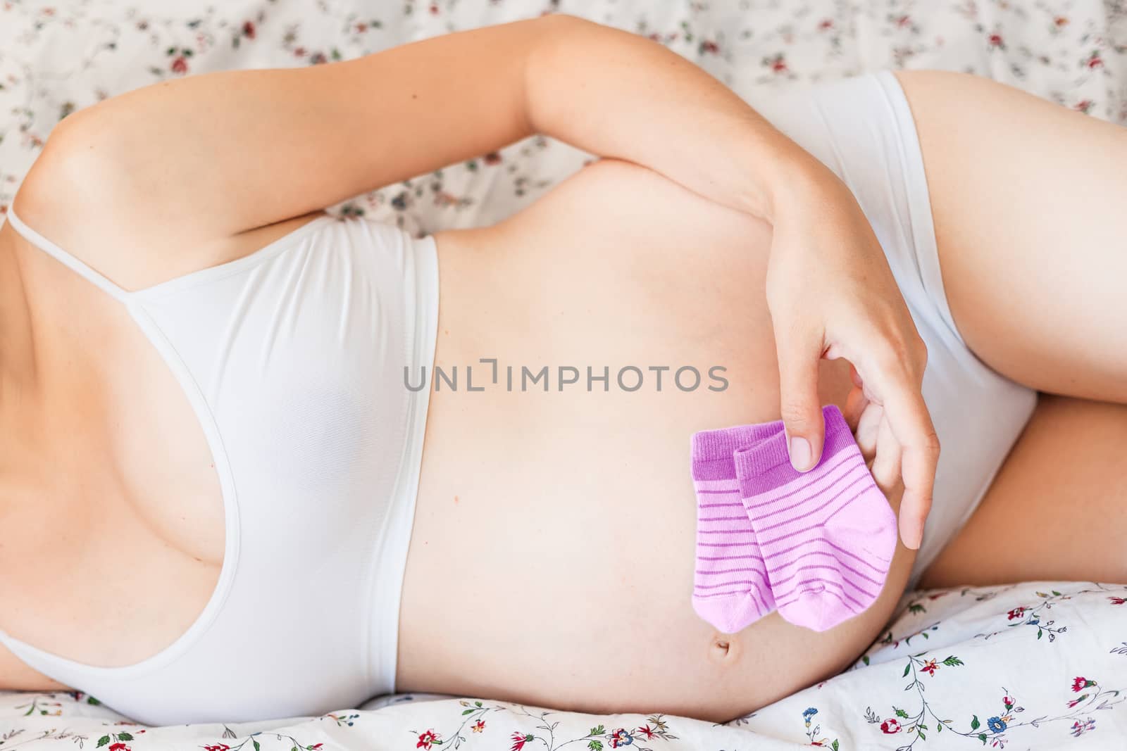 Pregnant woman in white underwear lying in bed with small baby's socks. Young woman expecting a baby girl.