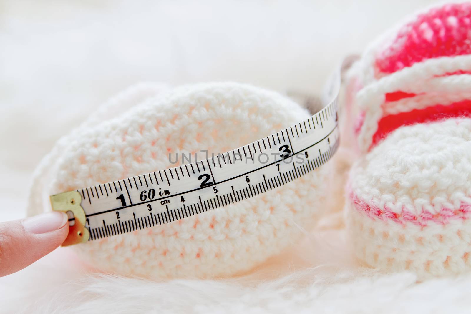 Little baby shoes. Hand knitted first sneakers for newborn girl. Crochet handmade pink bootees on fluffy white background. Mother measures the length of booties.