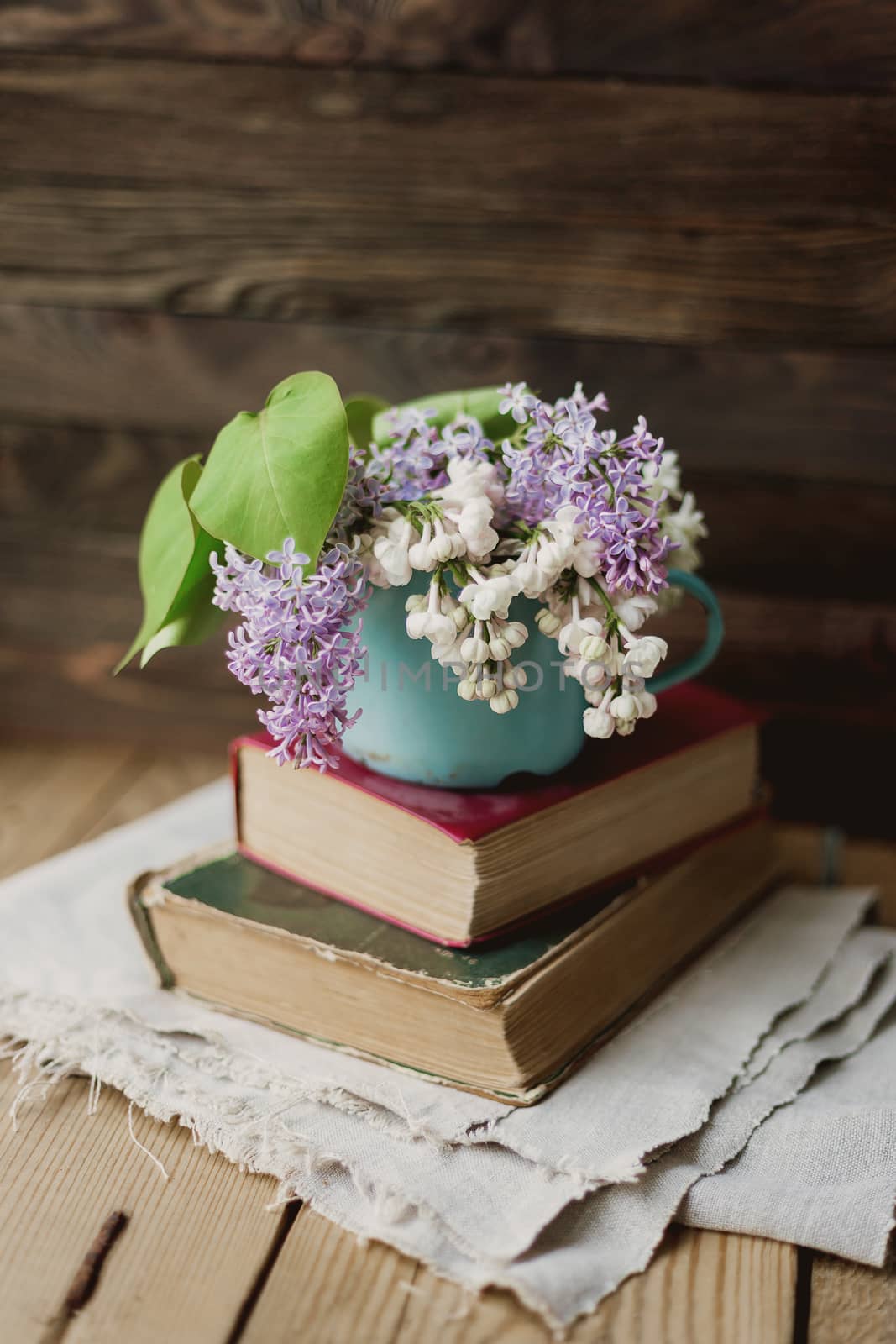 Two old books and rusty mug with lilac bouquet on homespun napkin. Interesting reading, vintage background.