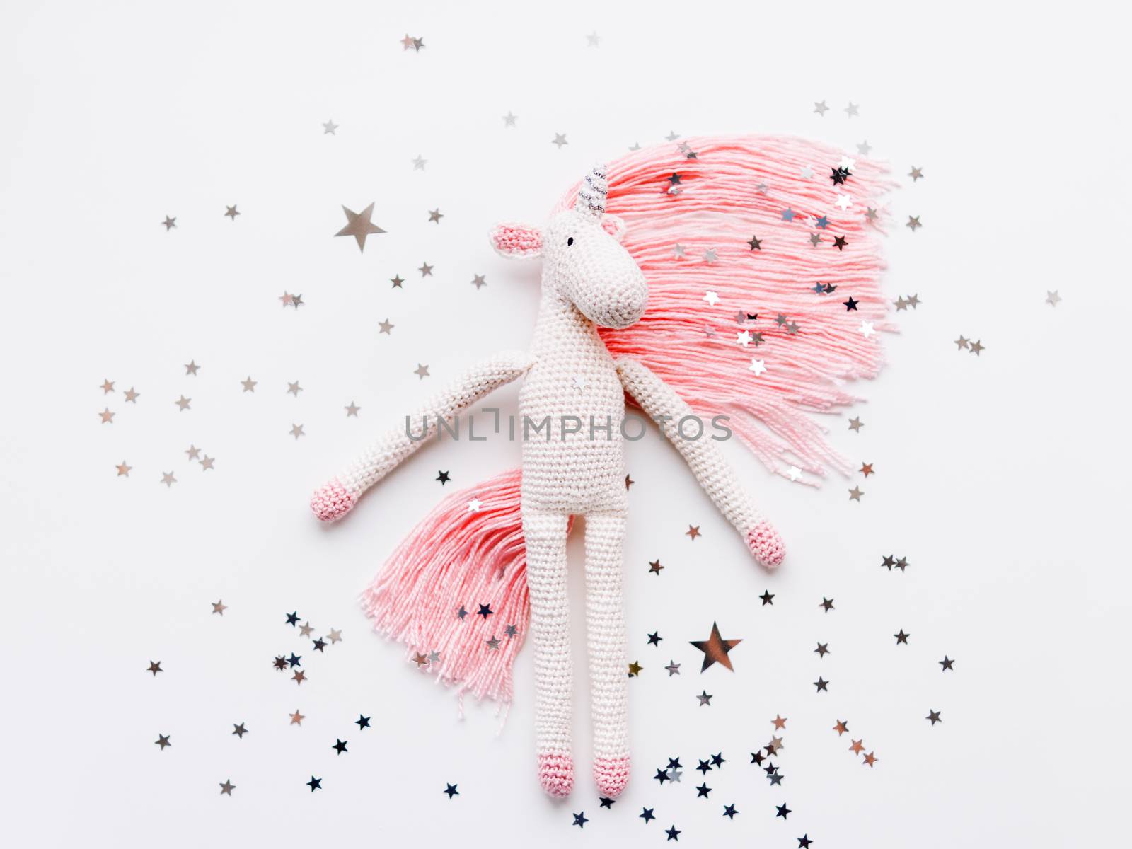 Cute fairy unicorn with a pink mane and a tail made of threads. Crocheted hand made toy on white background with silver stars confetti. Trendy creature, symbol of magic and miracles.