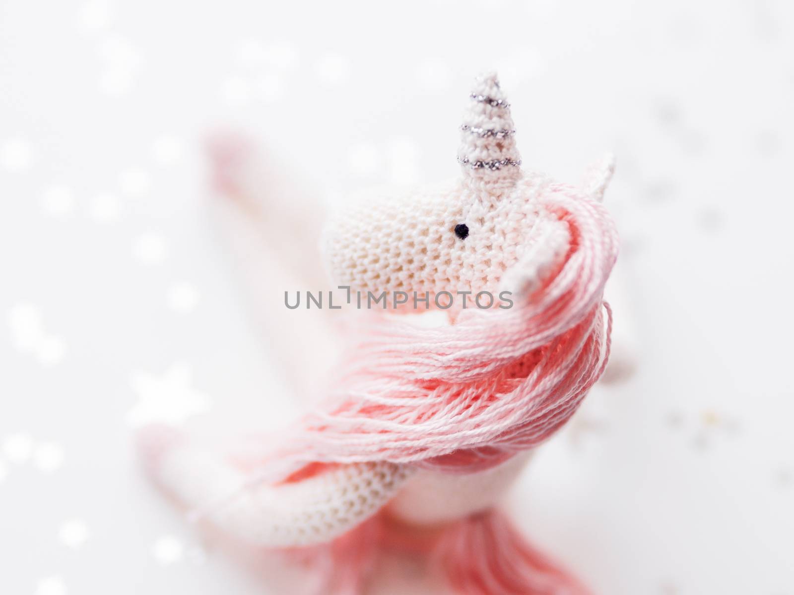 Cute fairy unicorn with a pink mane and a tail made of threads. Crocheted hand made toy on white background with silver stars confetti. Trendy creature, symbol of magic and miracles.