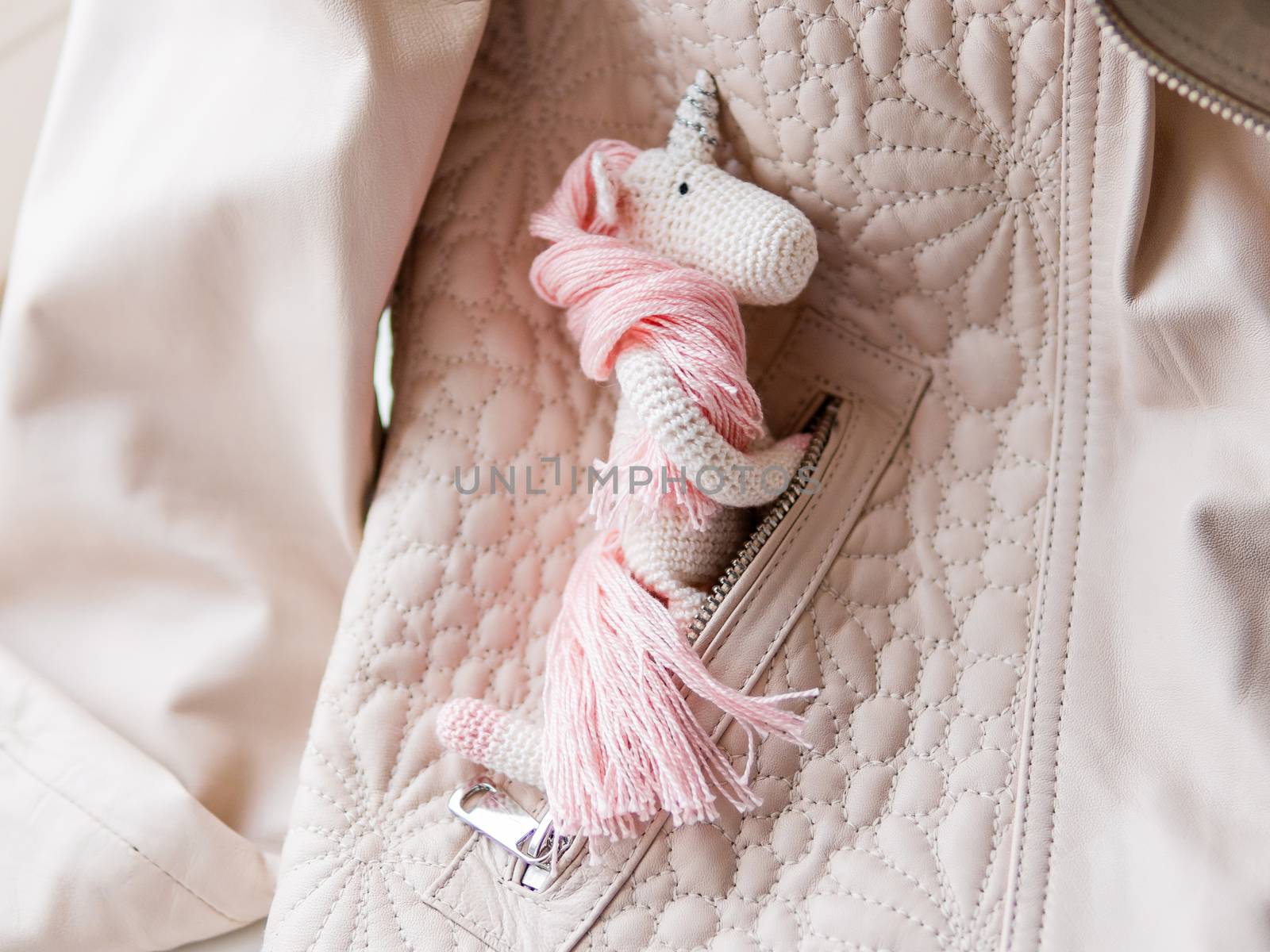 Cute fairy unicorn with a pink mane and a tail made of threads. Crocheted hand made toy in pocket of leather jacket. Trendy creature, symbol of magic and miracles.