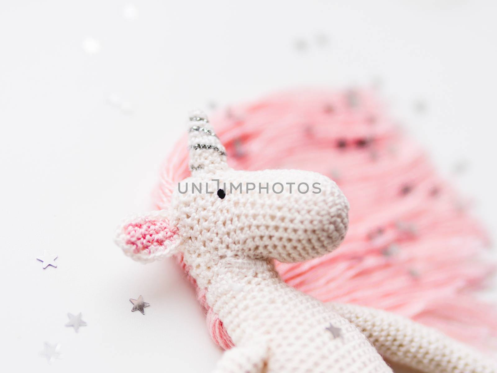 Cute fairy unicorn with a pink mane and a tail made of threads. Crocheted hand made toy on white background with silver stars confetti. Trendy creature, symbol of magic and miracles.