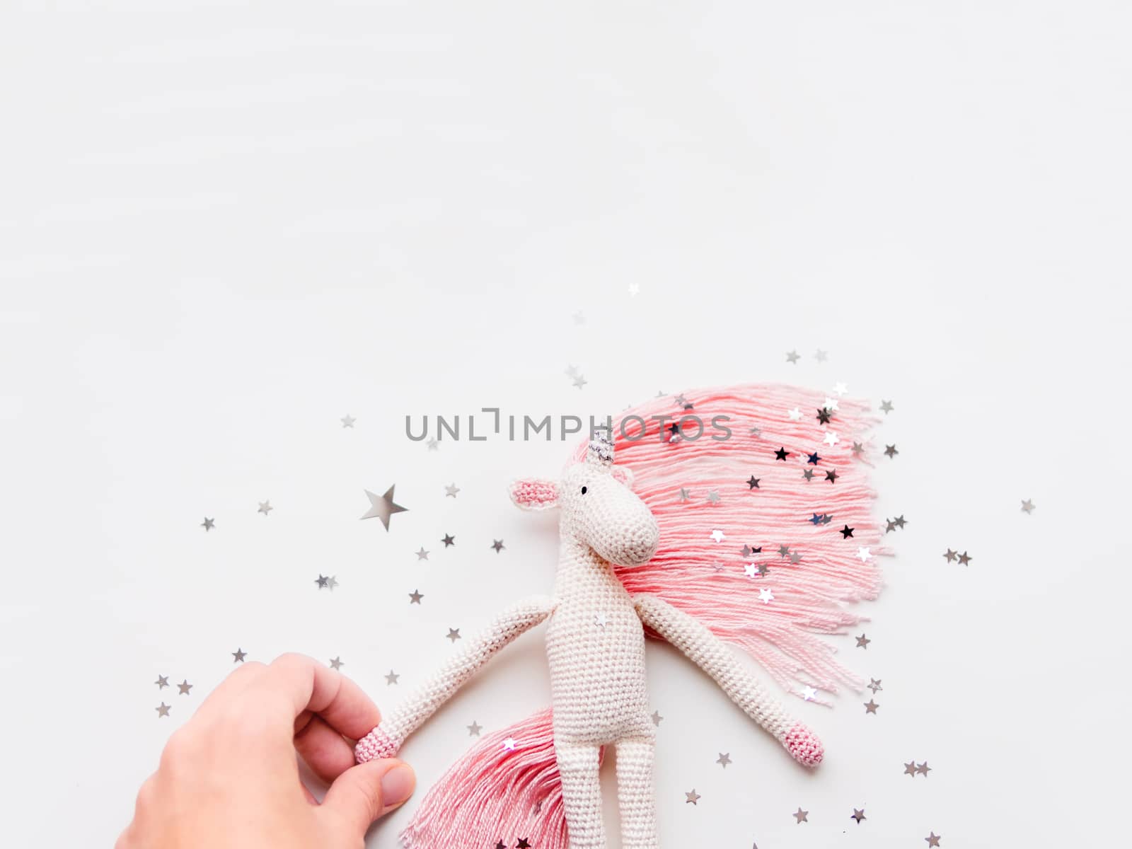 Cute fairy unicorn with a pink mane and a tail made of threads. Crocheted hand made toy on white background with silver stars confetti. Trendy creature, symbol of magic and miracles.