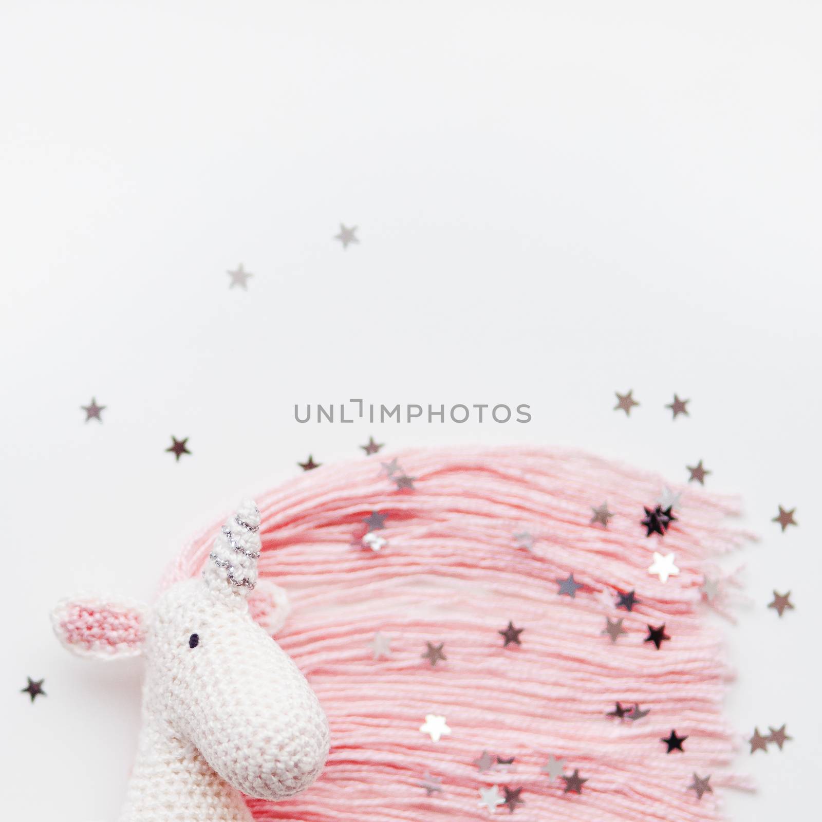 Cute fairy unicorn with a pink mane and a tail made of threads. Crocheted hand made toy on white background with silver stars confetti. Trendy creature, symbol of magic and miracles. Place for text. by aksenovko