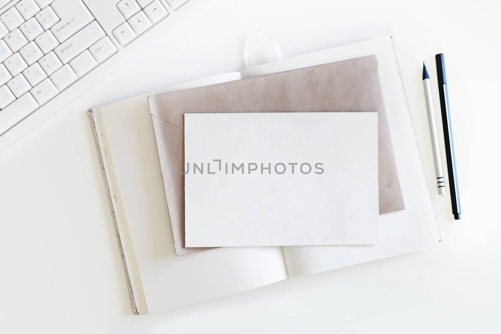 Top view on white table with keyboard, copybook with blank pages, craft envelope, clear postcard and pen with pencil. Flat lay.