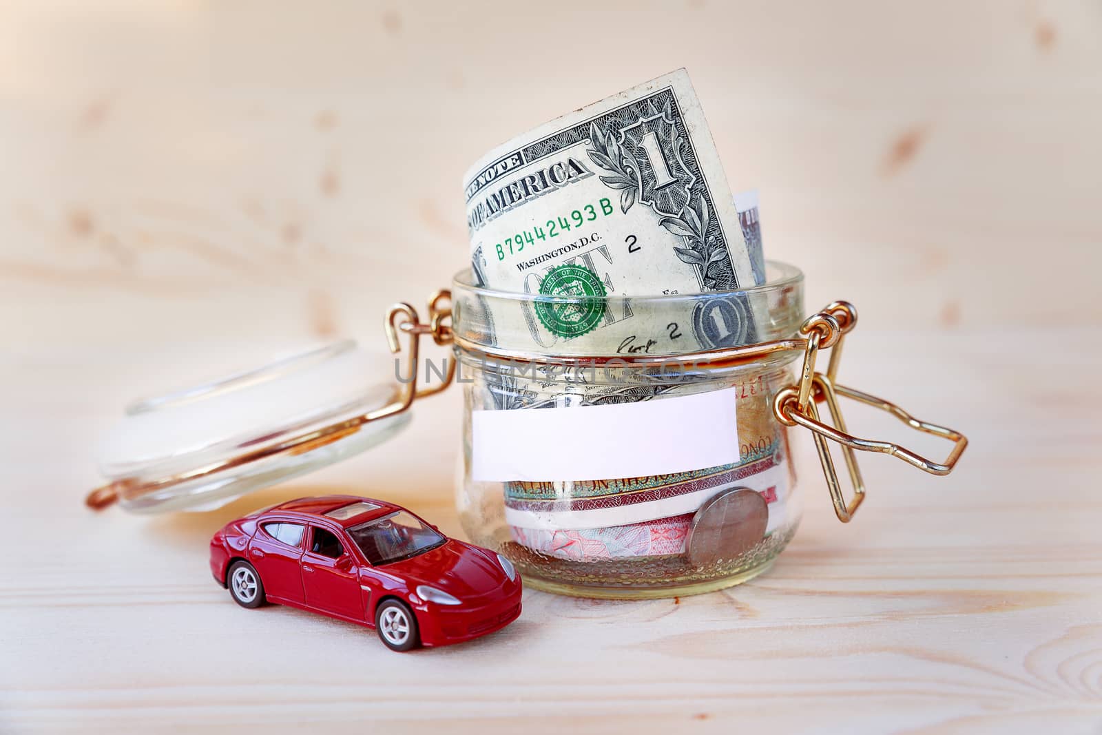 Collecting money for new car. Glass tin as moneybox with cash savings (banknotes and coins) and toy car on wooden background. White empty space for your text.