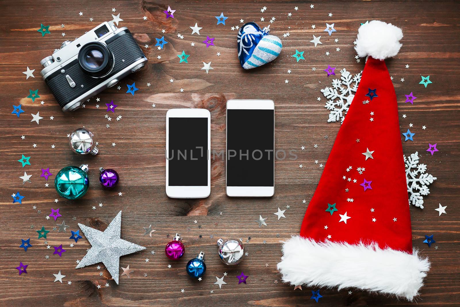 Christmas and New Year background with two smartphones, old fashioned camera, red Santa's hat, decorations - balls, stars, silver sparkling snowflakes, confetti on wooden table. Mock up, place for your text.