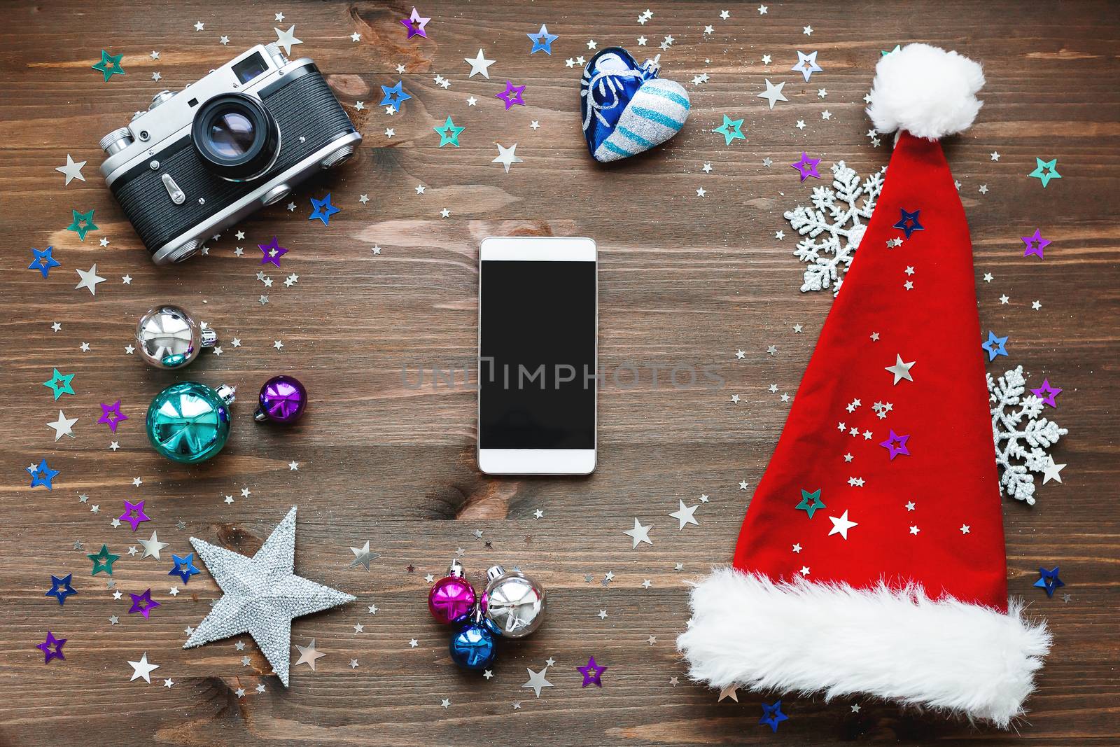 Christmas and New Year background with smartphone, old fashioned camera, red Santa's hat, decorations - balls, stars, silver sparkling snowflakes, confetti on wooden table. Mock up, place for your text.