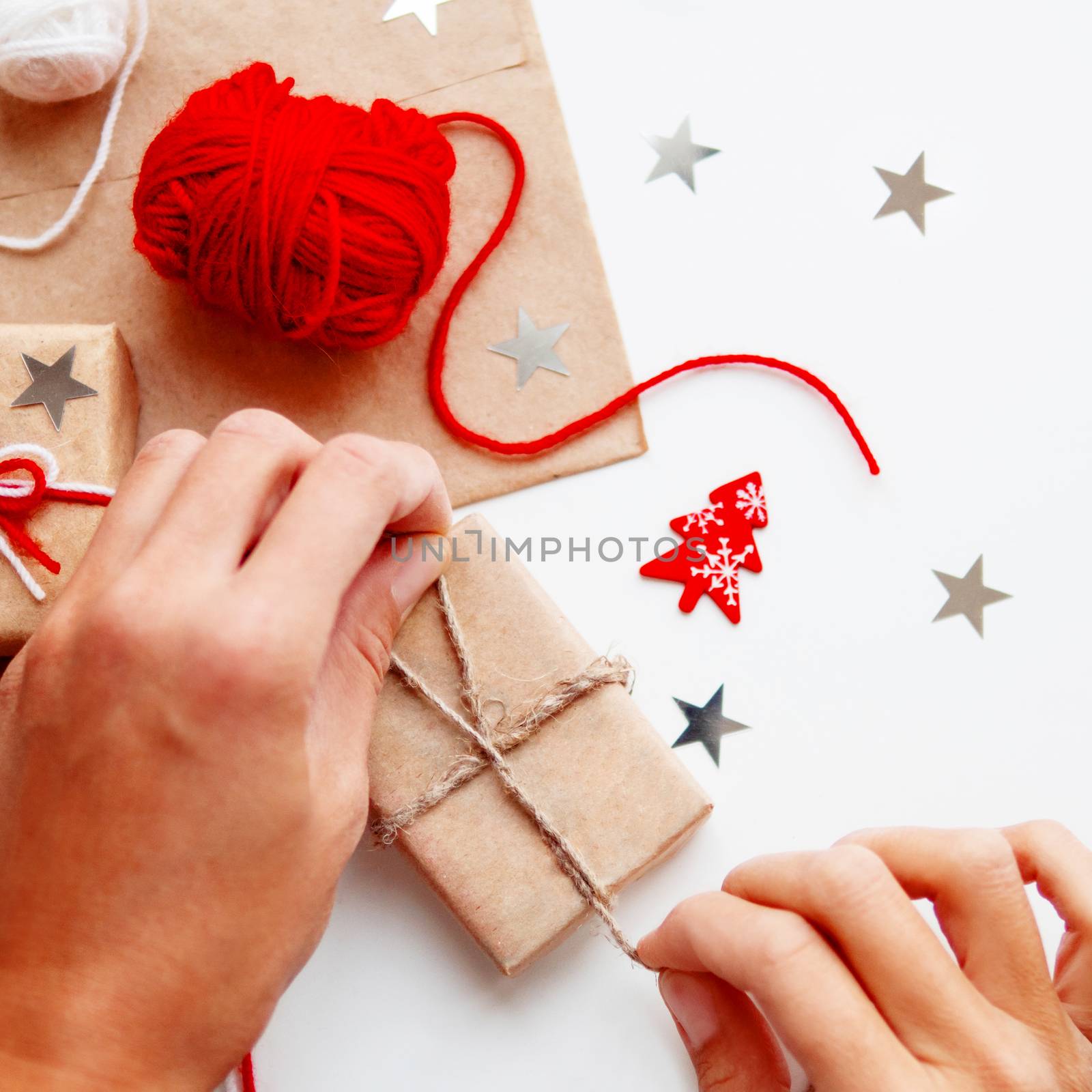 Woman is packing Christmas and New Year DIY presents in craft paper. Gifts tied with white and red threads. Boxes and star confetti on white background.