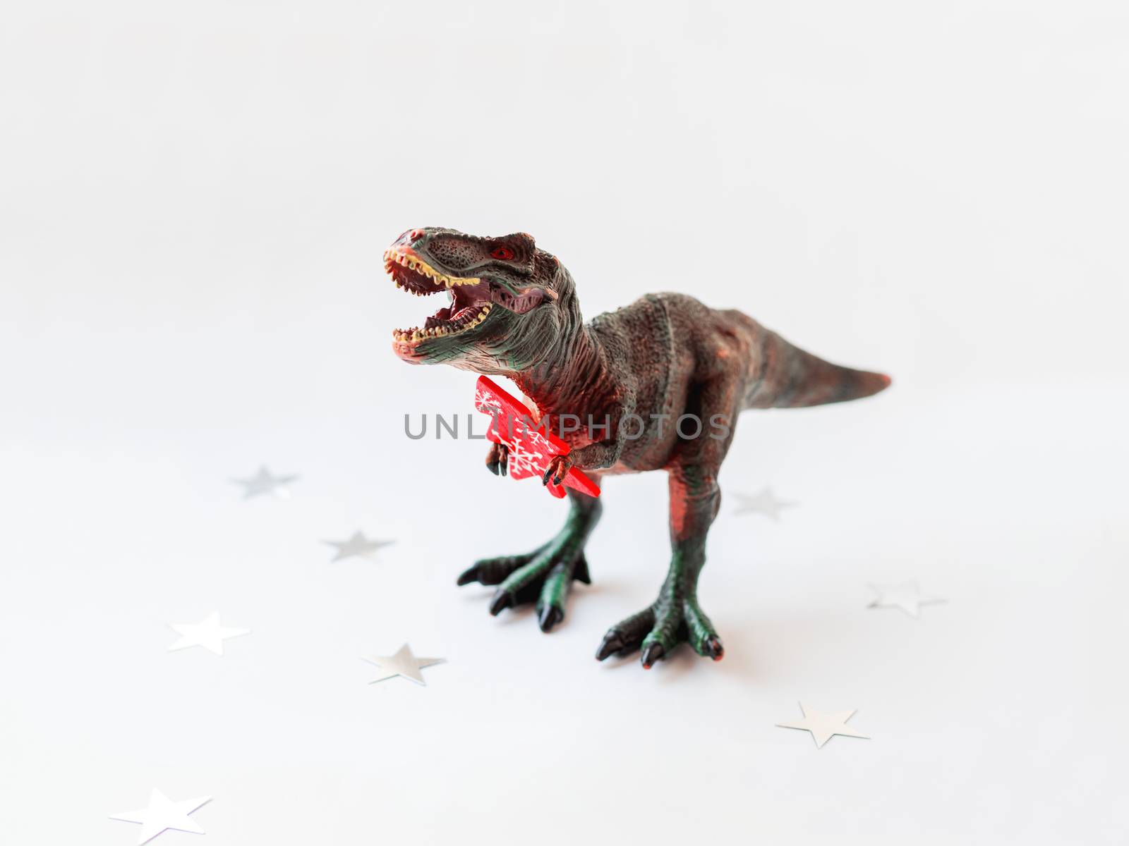 Dinosaur with red Christmas tree. Plastic toy with wooden symbol of Christmas and New Year. White background with star confetti.
