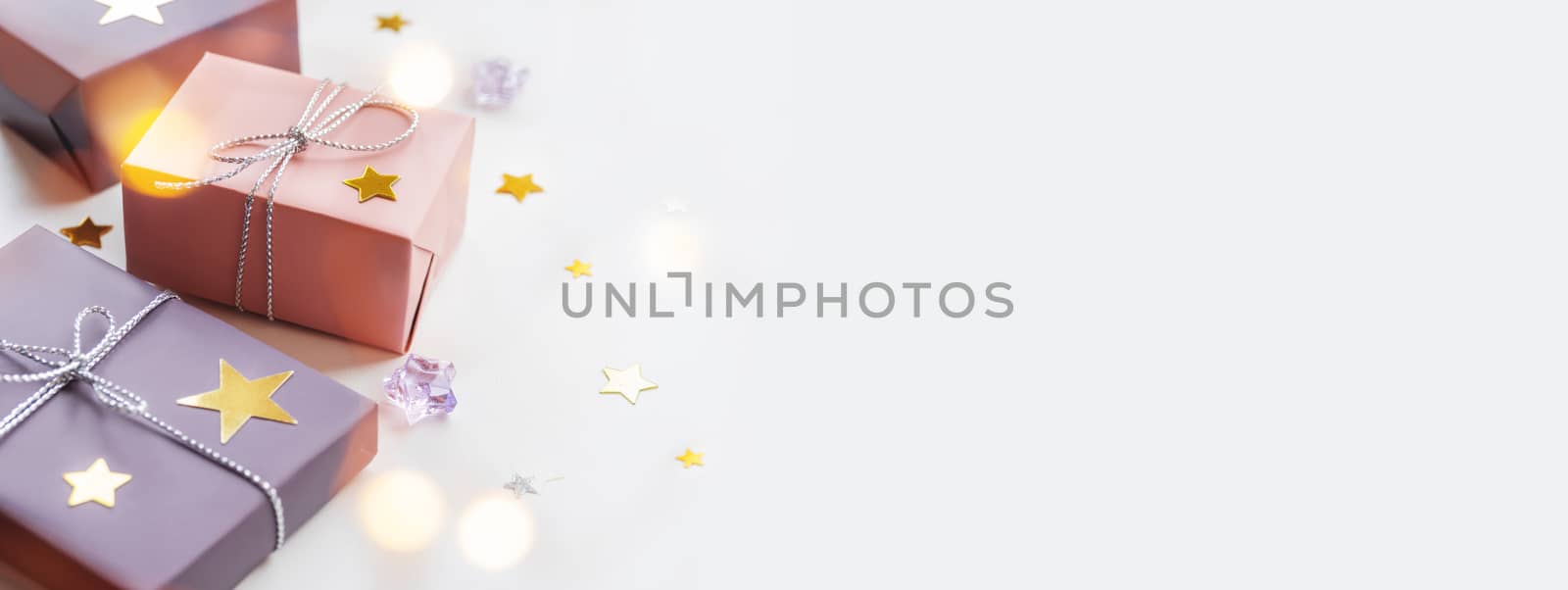 Holiday presents. Gifts wrapped in pale pink and violet paper with silver ribbons and bow. Stars confetti, light bulbs bokeh and white copy space.