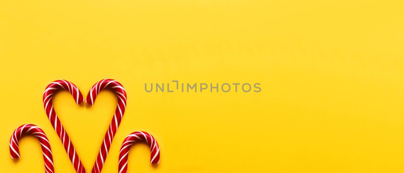 Heart made of two Christmas candy cones on yellow background. Colorful holiday sweets with bright copy space. Traditional dessert wtih red and white stripes.