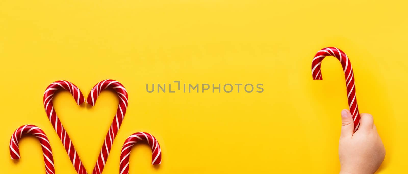 Heart made of two Christmas candy cones on yellow background. Colorful holiday sweets with bright copy space. Traditional dessert wtih red and white stripes.