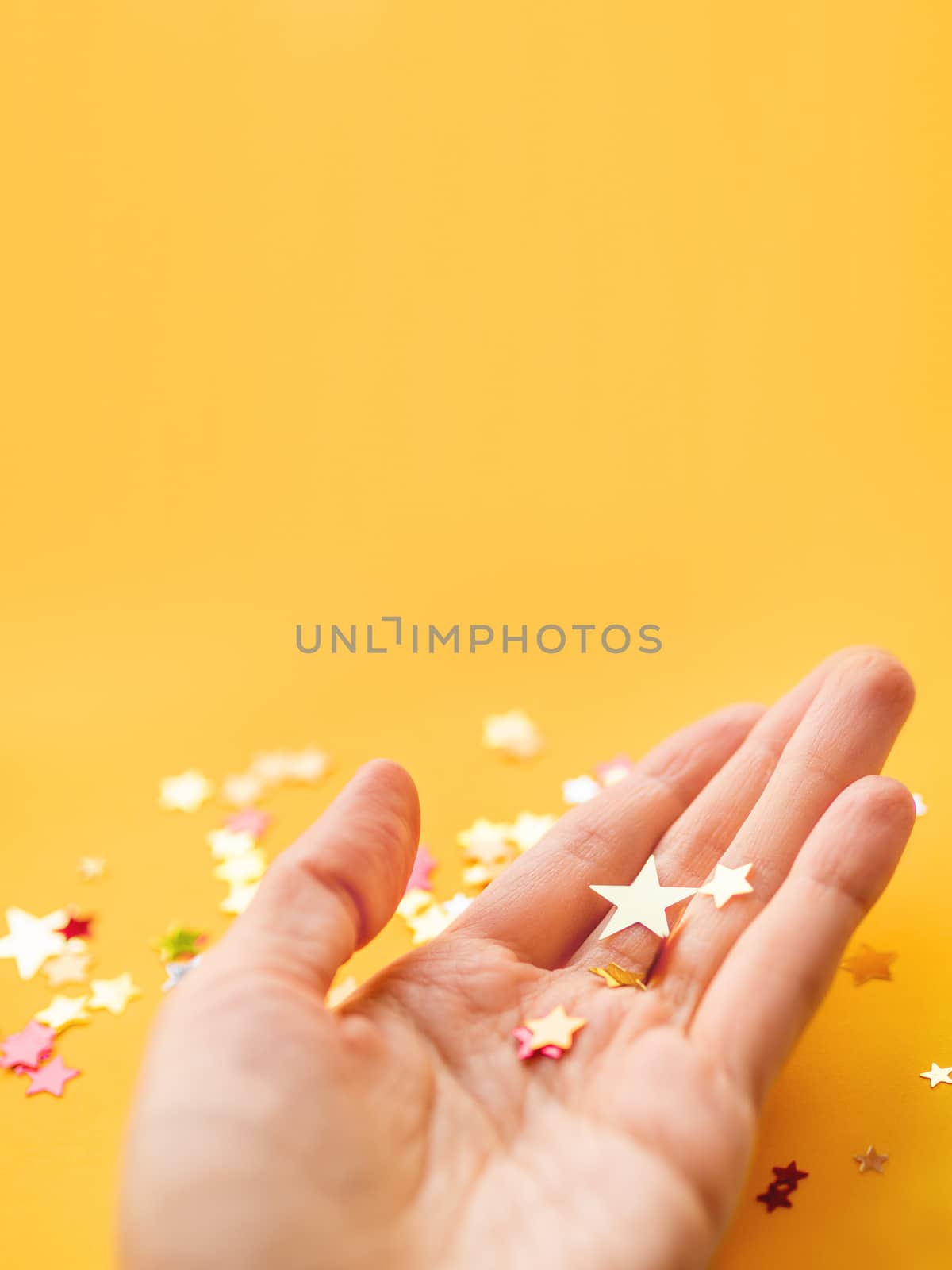 Yellow holiday background with colorful star confetti on hand. Copy space for Christmas and New Year cards.