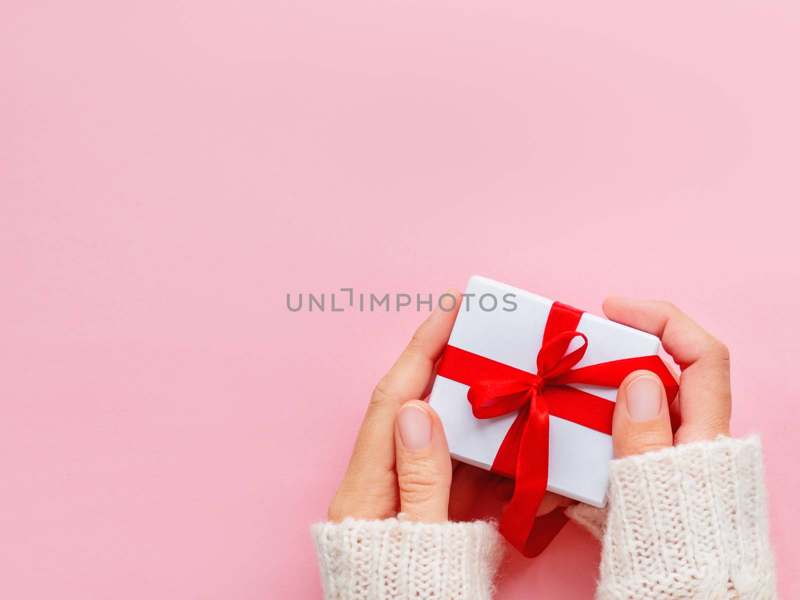 Woman holds a present in white wrapping paper on pink backdrop. Holiday gift with red ribbon and bow. Copy space.