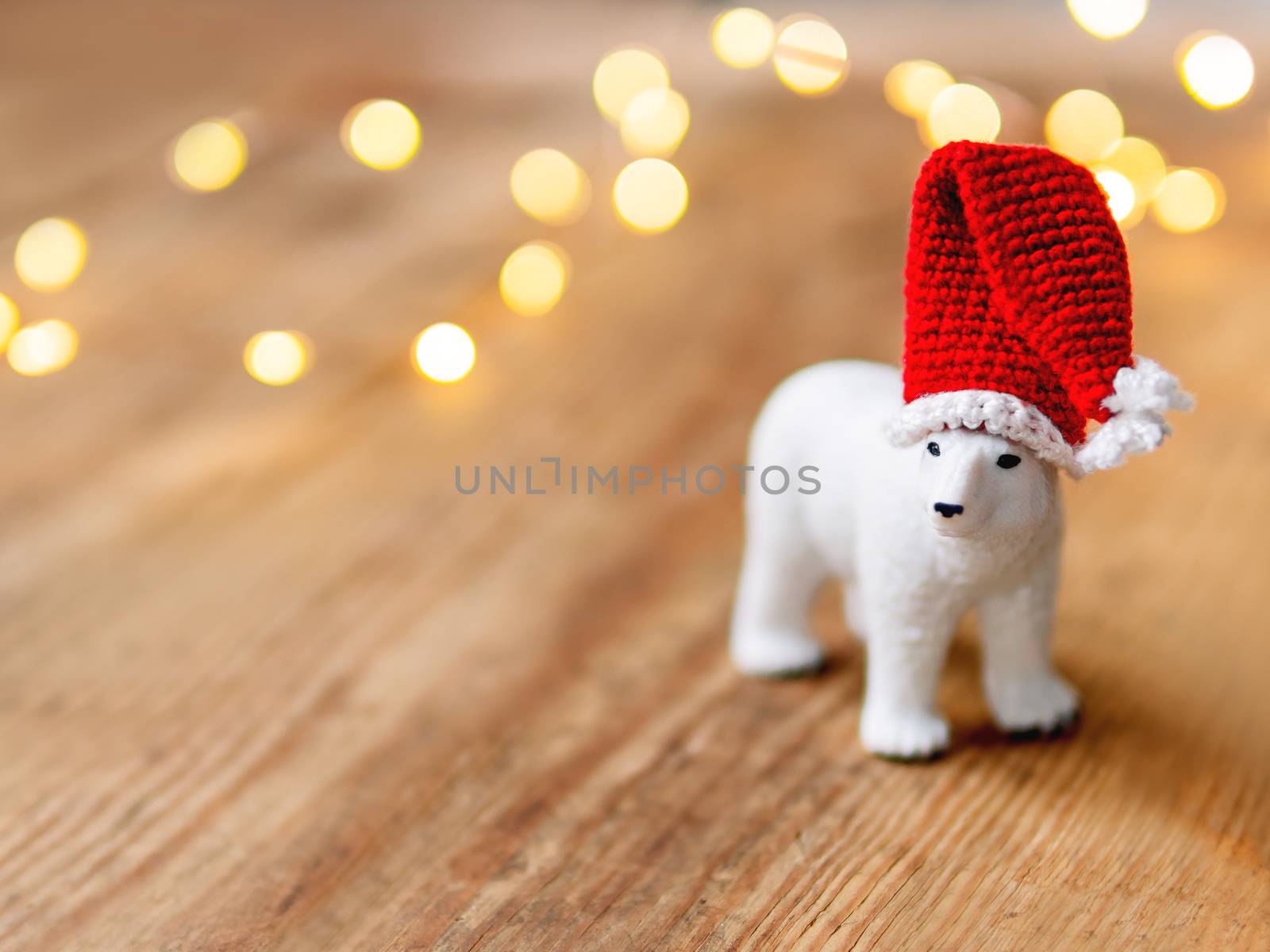 Cute polar bear in Santa's hat standing on shabby wooden table with light bulbs on background.