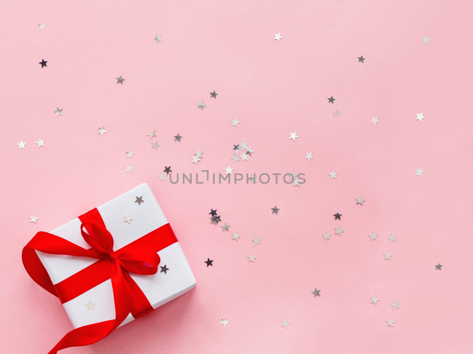 Present in white wrapping paper on pink backdrop with silver star confetti. Holiday gift with red ribbon and bow. Top view, flat lay.