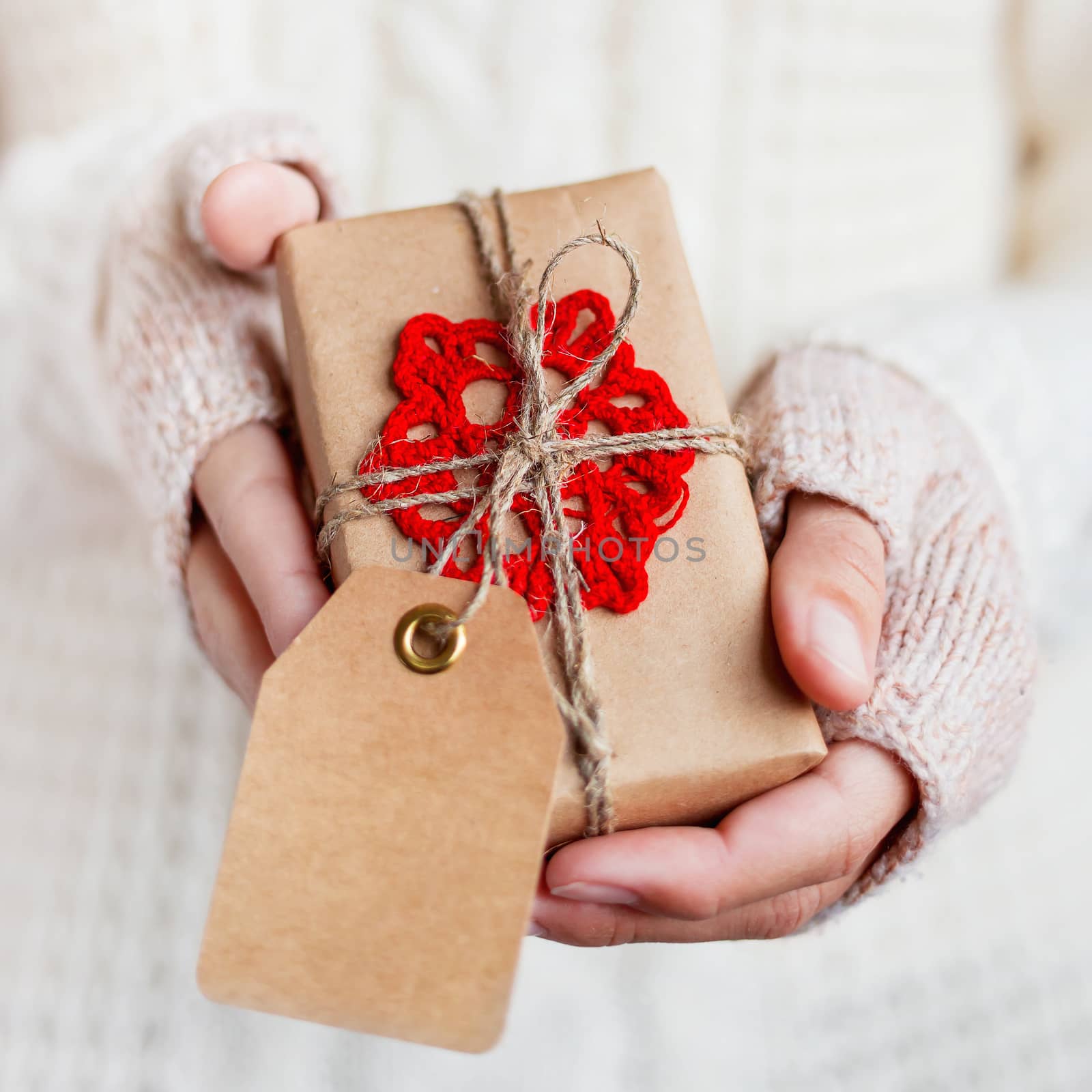 Woman in white knitted sweater and mitts holding a present packed in craft paper with crocheted red snowflake. DIY Christmas or New Year, St. Valentine Day gift with tag.