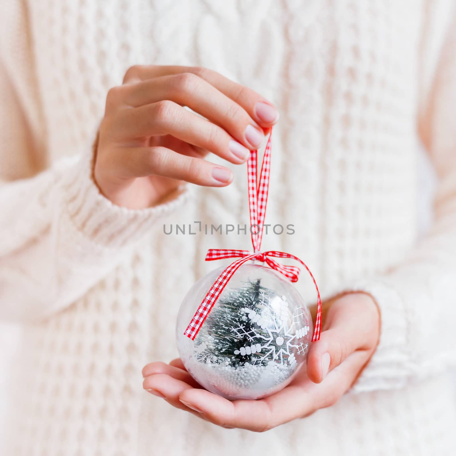 Woman in knitted sweater is holding Christmas decoration - transparent glass ball with ribbon and snowy fir tree inside. New Year snow globe.