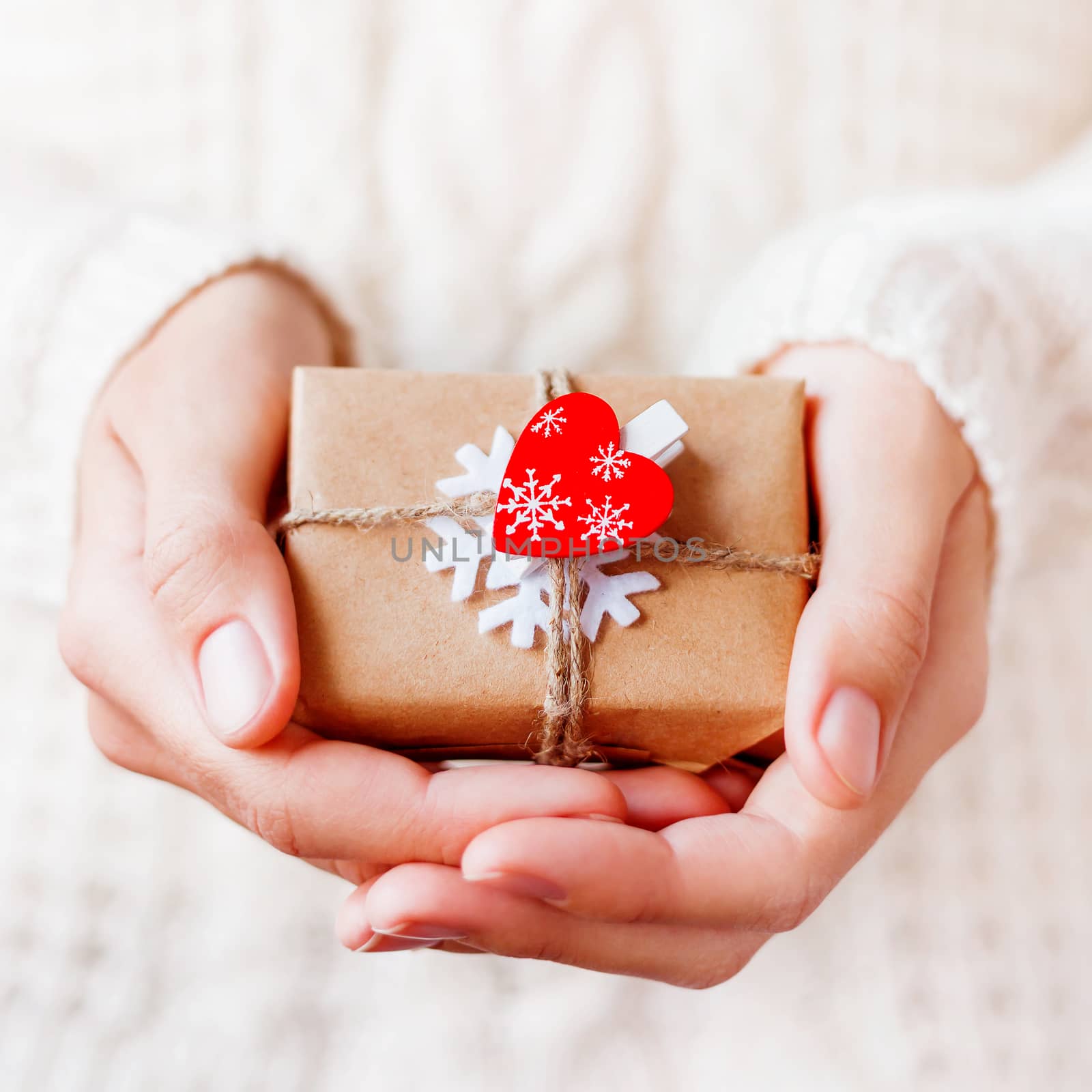 Woman in knitted sweater is holding Christmas present packed in craft paper with felt snowflake and red heart. New Year or St. Valentine Day DIY gift.