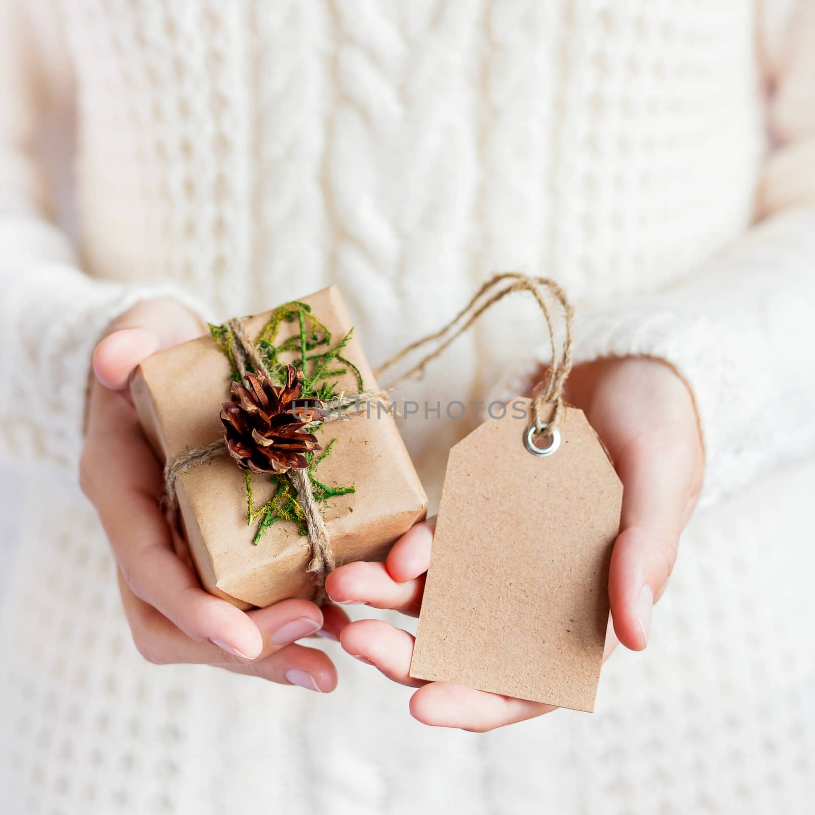 Woman in white knitted sweater is holding a present packed in craft paper with pine cona and tag. DIY Christmas or New Year, St. Valentine Day gift.