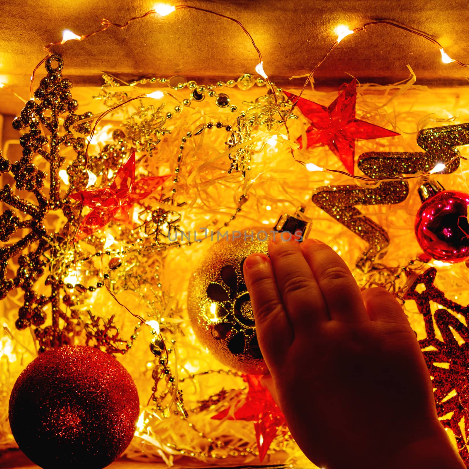 Cardboard box with Christmas and New Year decorations and light bulbs. Little baby touching golden sparkling ball with flower.