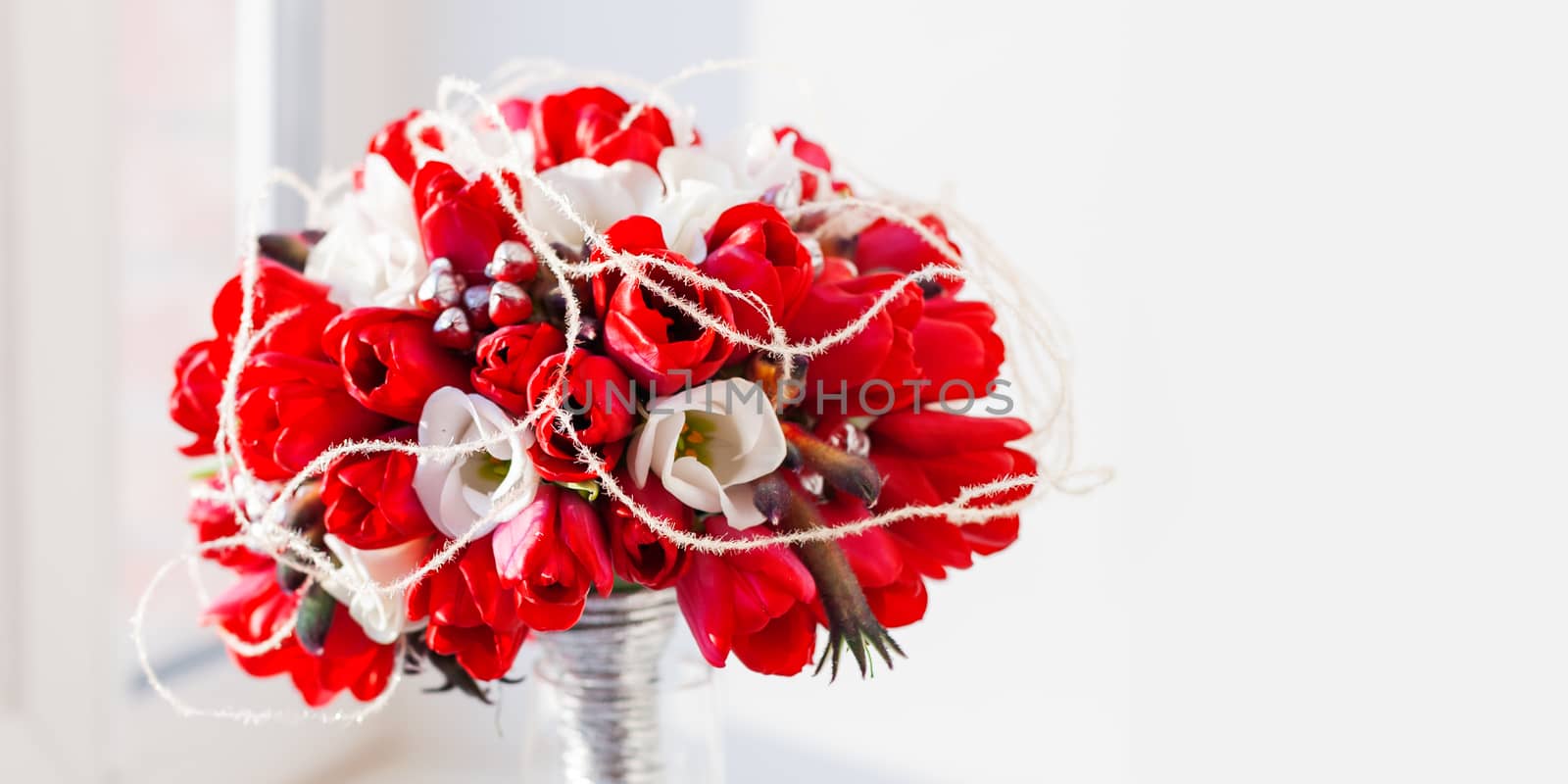 Bridal bouquet in glass vase on sunlight. Traditional floral composition with bright red tulips on window sill. Background with copy space. by aksenovko