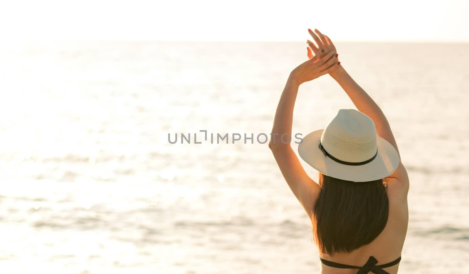 Back view of happy young Asian woman in black bikini and straw hat relax and enjoy at tropical paradise beach at sunset. Girl in summer vacation fashion. Sexy model. Elegant lifestyle. Summer vibes.