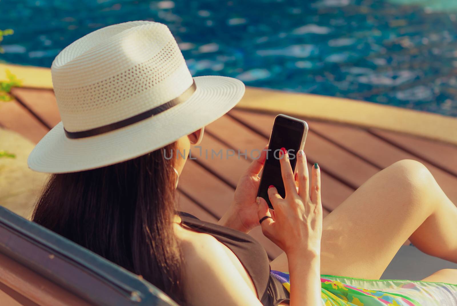 Asian woman with hat and swimsuit sitting in chair at poolside and using smartphone on summer vacation. Luxury life. Checking News on mobile phone application by swimming pool. Girl with nail manicure