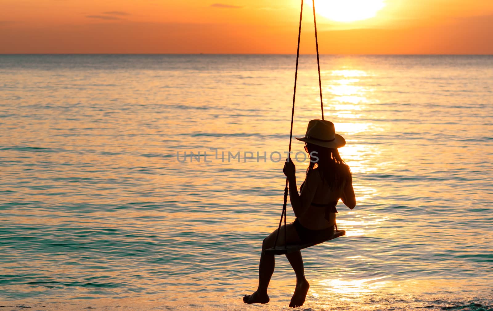Silhouette woman wear bikini and straw hat swing the swings at the beach on summer vacation at sunset. Enjoying and relaxing girl on holiday. Summer vibes. Woman watching beautiful sunset sky. 