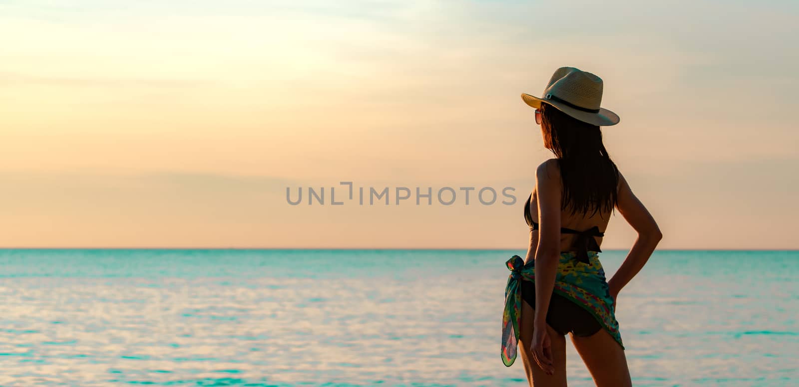 Back view of happy young Asian woman in black swimsuit and straw hat relax and enjoy holiday at tropical paradise beach at sunset. Girl in summer vacation fashion. Beauty sexy model. Elegant lifestyle