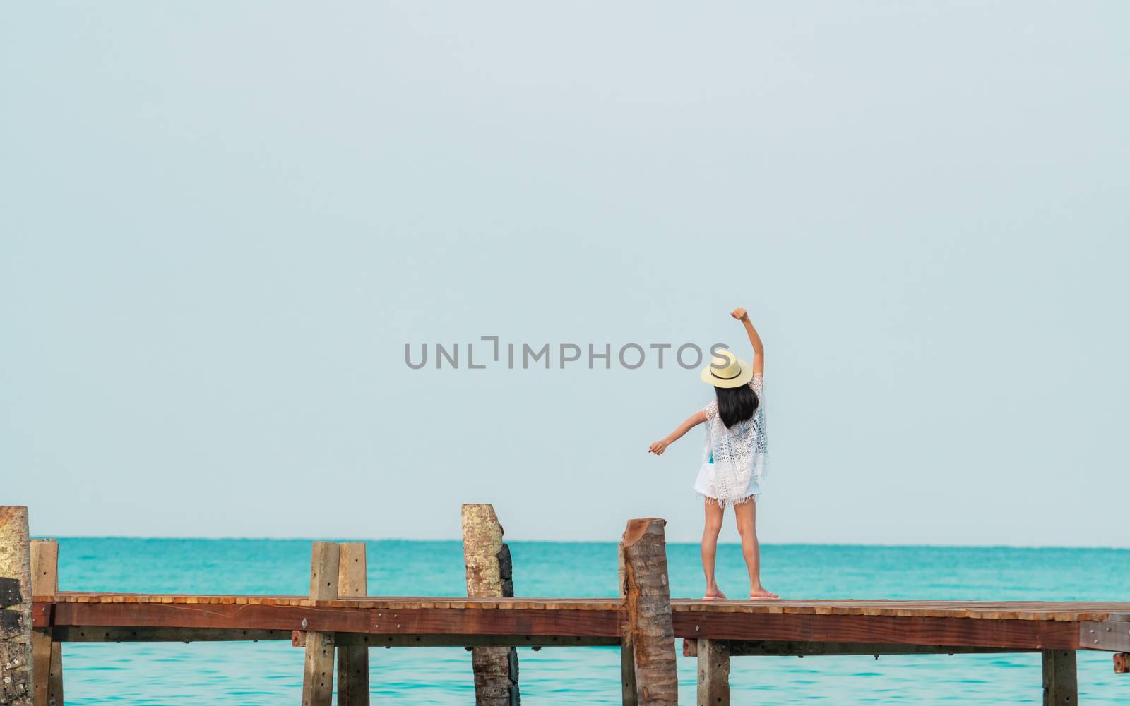 Back view of happy young Asian woman in casual style fashion and straw hat relax and enjoy holiday at tropical paradise beach. Girl stand at the wood pier of resort in summer vacation. Summer vibes.