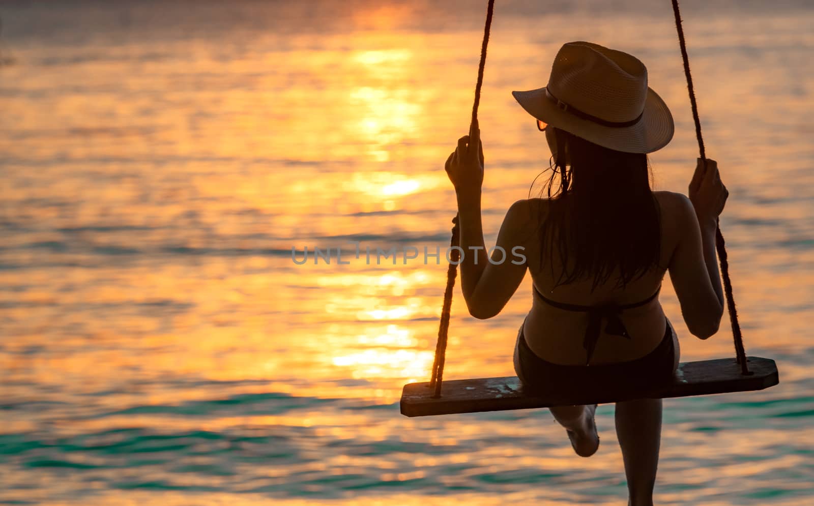Silhouette woman wear bikini and straw hat swing the swings at the beach on summer vacation at sunset. Enjoying and relaxing girl on holiday. Summer vibes. Woman watching beautiful sunset sky. 