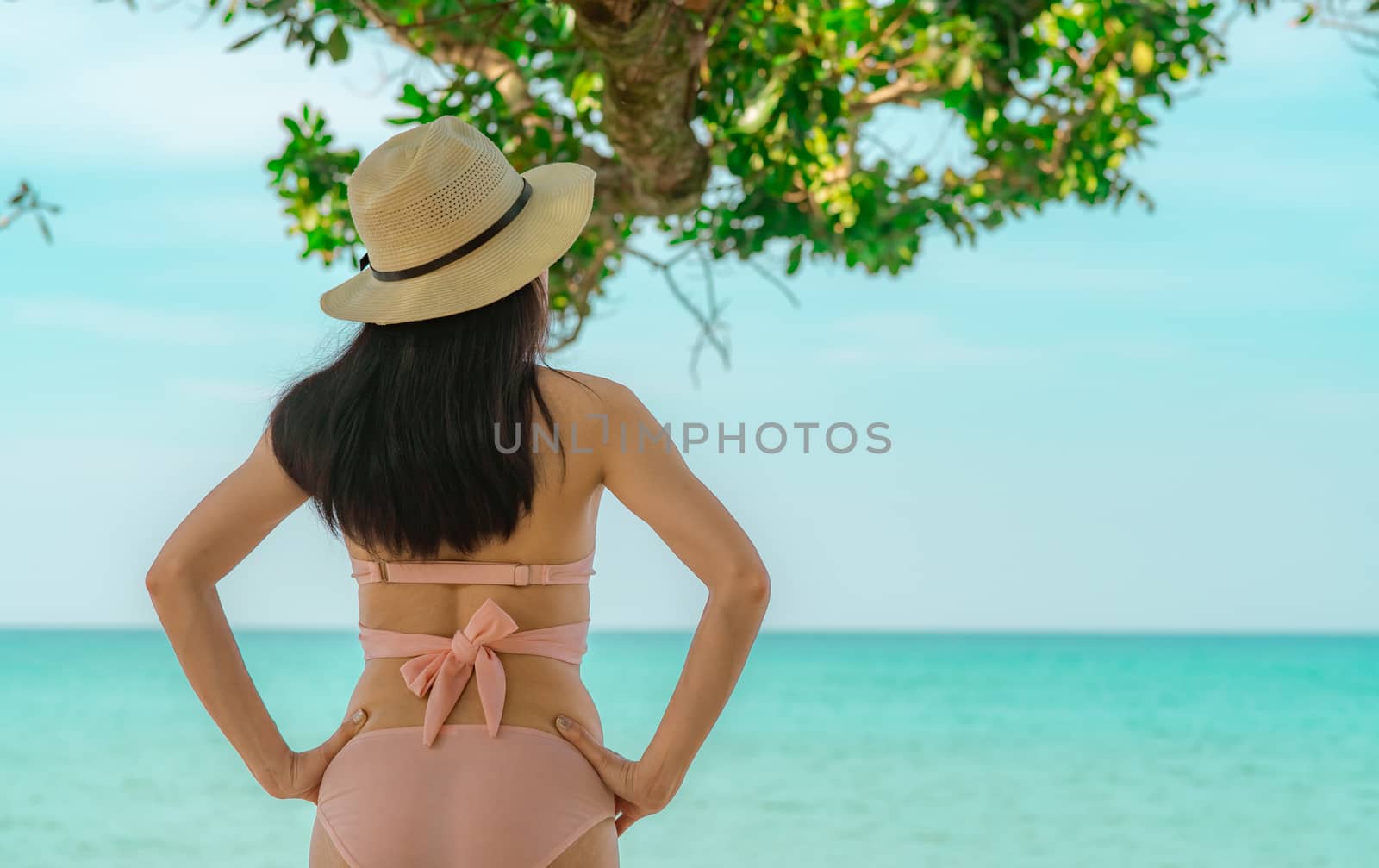 Back view of happy young Asian woman in pink swimsuit and straw hat relaxing and enjoy holiday at tropical paradise beach on vacation. Girl in summer vacation fashion. Beauty sexy model. Summer vibes.